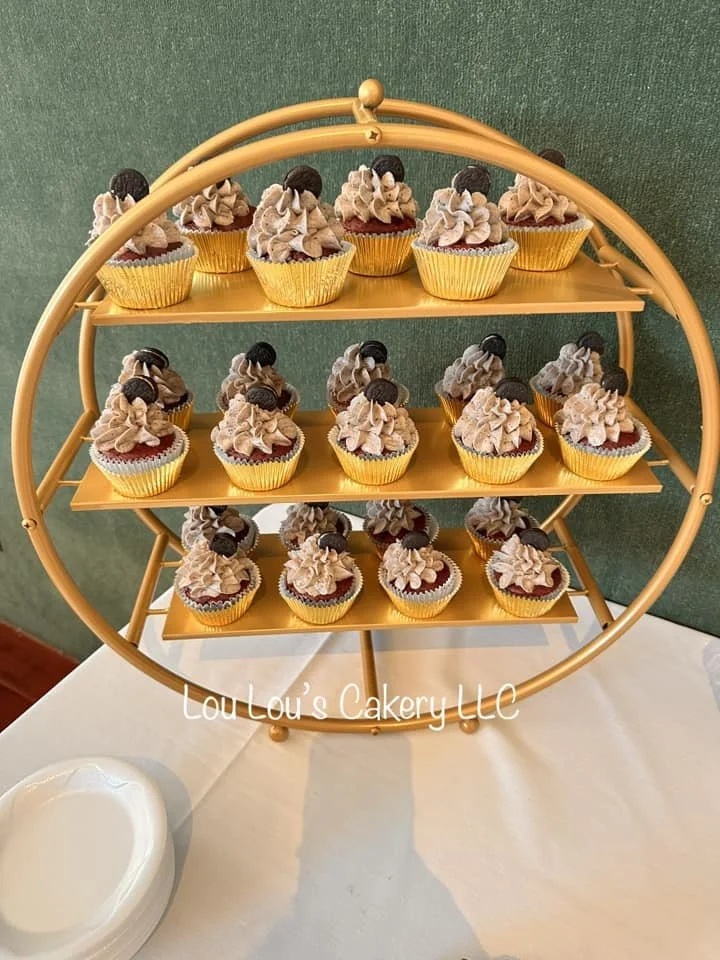 A three-tiered golden cupcake stand displaying multiple chocolate cupcakes with beige frosting topped with Oreo cookies, set on a white table with a small white bowl nearby. The background is green.