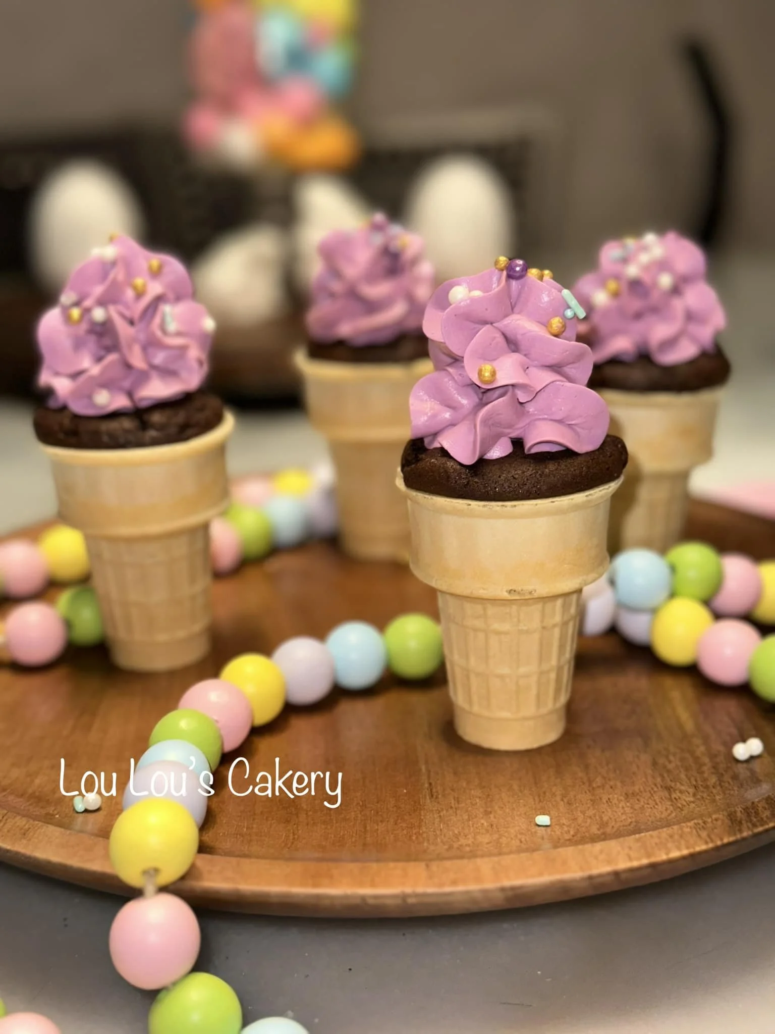 Decorative cupcakes designed to look like ice cream cones with pink frosting and sprinkles, displayed on a wooden board with pastel-colored bead garland.