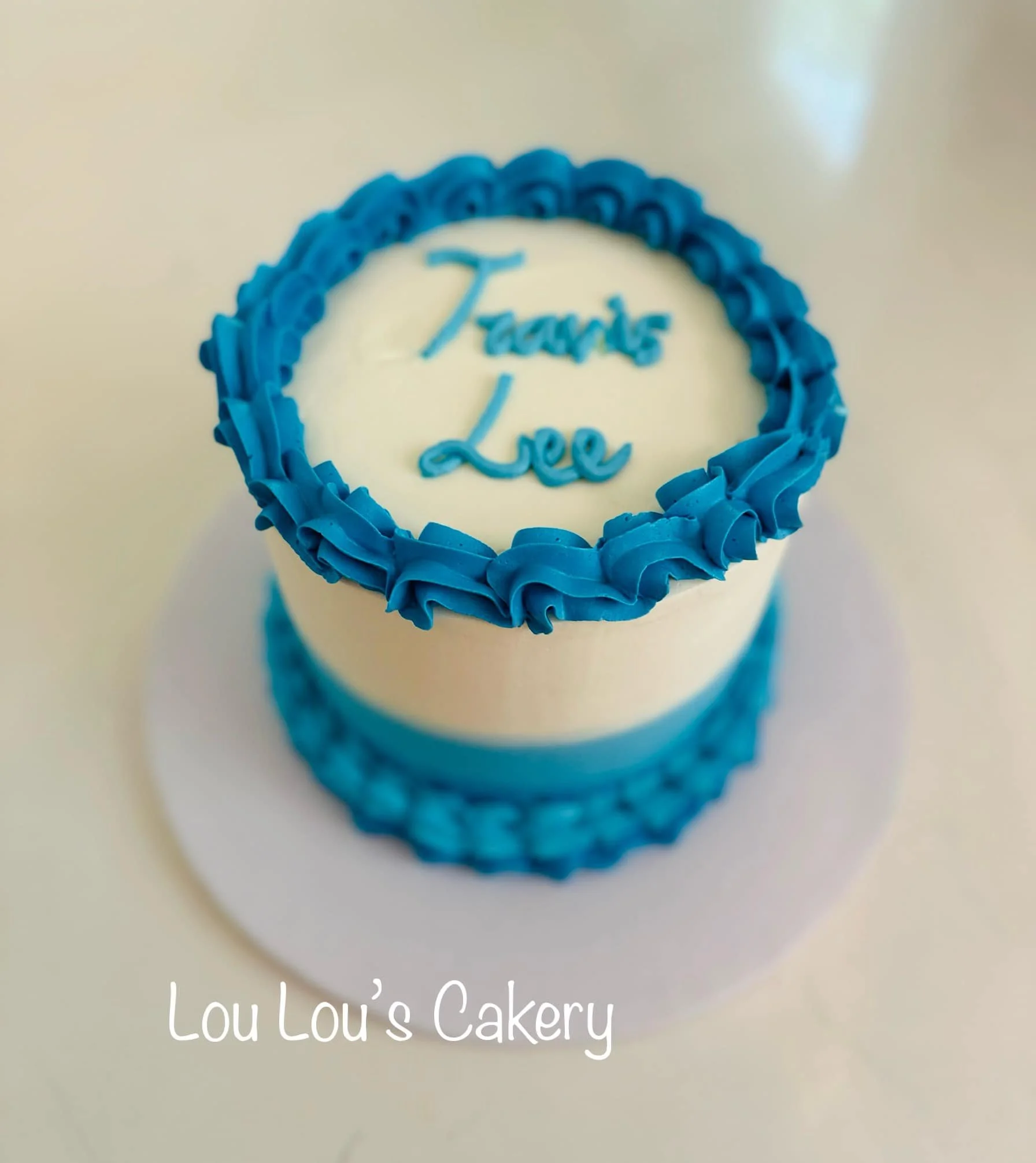 A white cake with blue icing decoration and writing that says 'Thanks Lee' on top, from Lou Lou's Cakery.