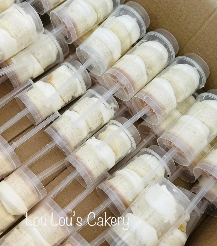 Small clear plastic cups filled with white cake and frosting, with a small clear plastic spoon inside each cup, arranged in a box. The text 'Lou Lou’s Cakery' is overlaid on the image.