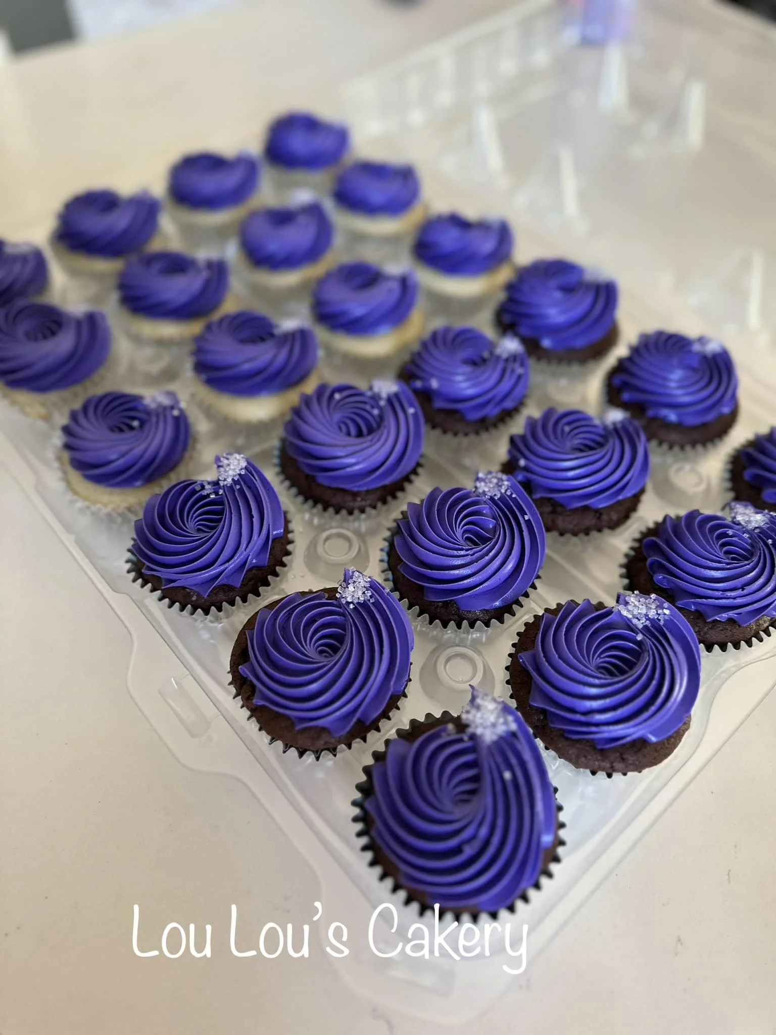 A tray of cupcakes with purple frosting and small white sugar decorations, labeled 'Lou Lou's Cakery'.