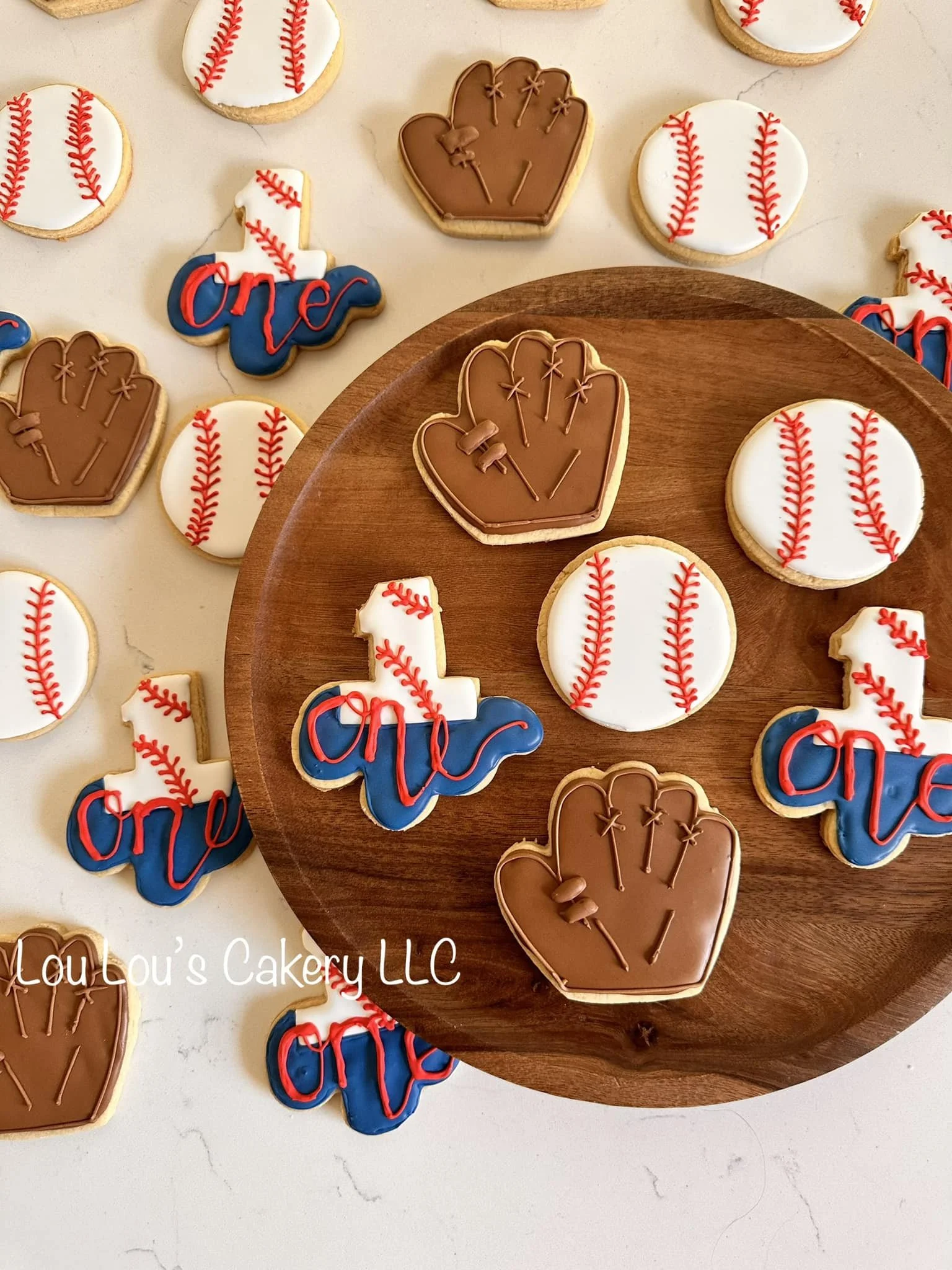 Cookies decorated with baseball and baseball glove designs, placed on a white surface and a wooden board. Some cookies spell out "one" with red and blue icing.