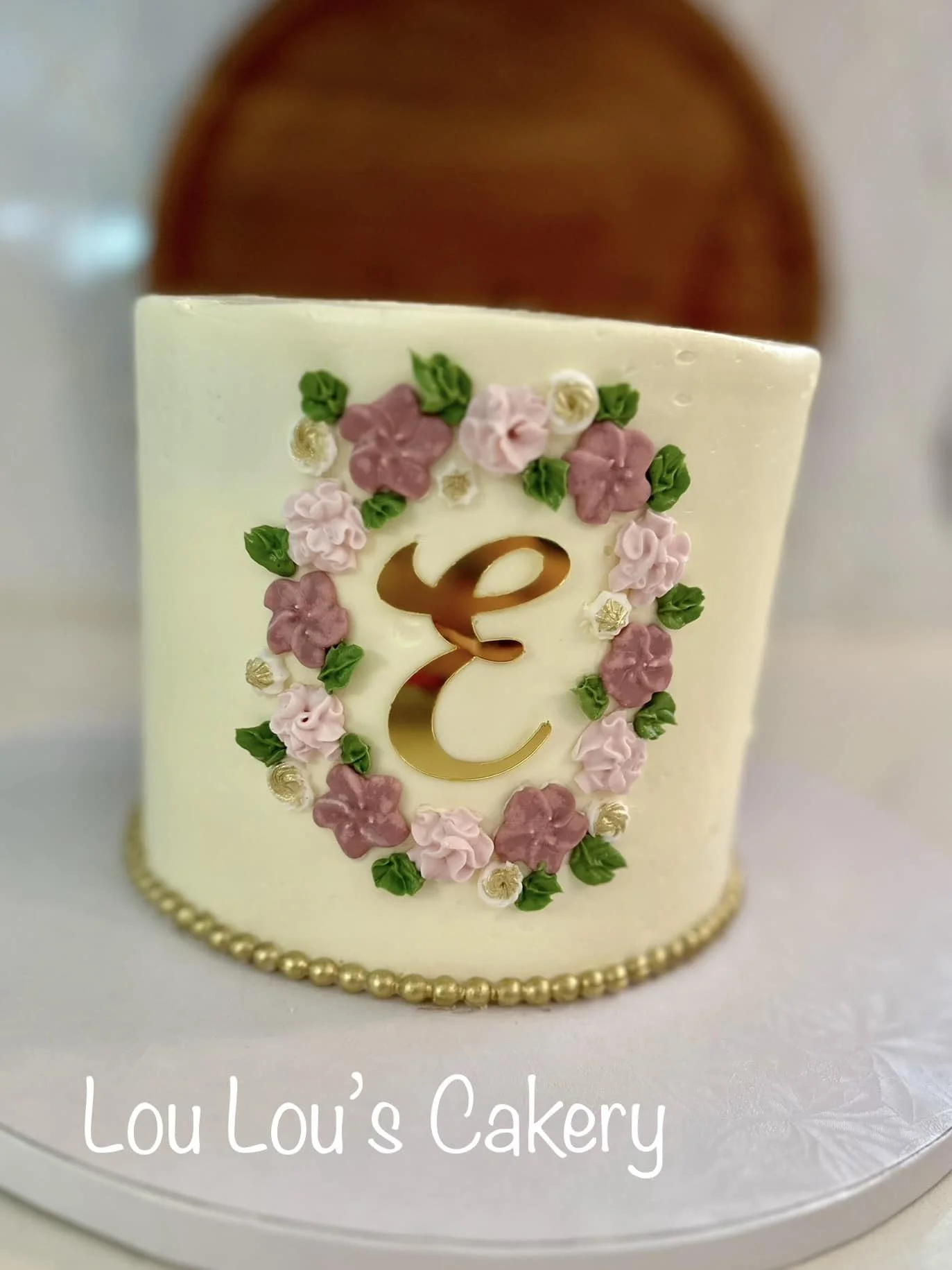 A white cake decorated with pink and white icing flowers, green leaves, and a gold letter 'E' in the center, with a border of gold beads at the bottom. The cake has the text 'Lou Lou's Cakery' at the bottom.