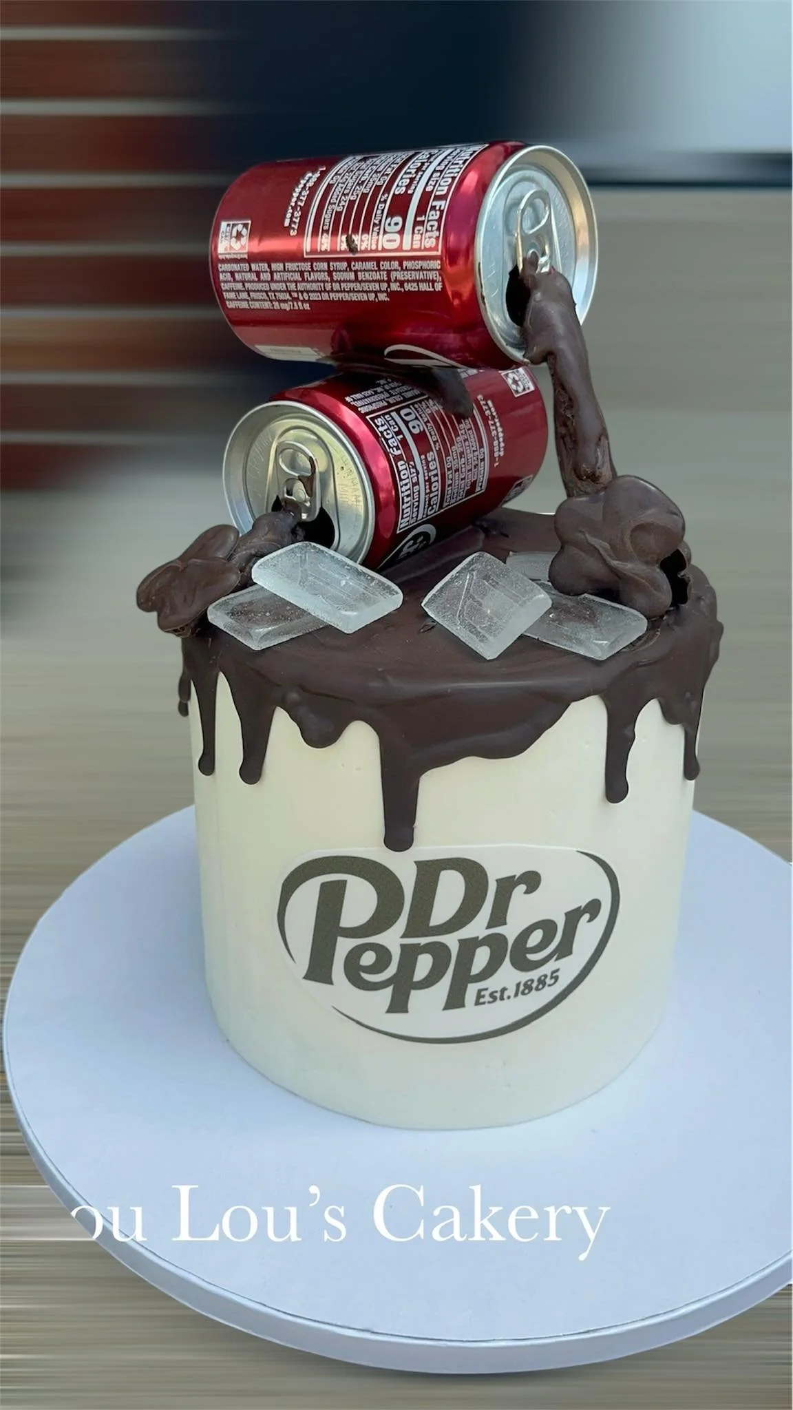 A cake decorated with a large Dr. Pepper logo, topped with two upside-down soda cans, chocolate icing, ice cubes, and chocolate decorations, sitting on a white cake stand.