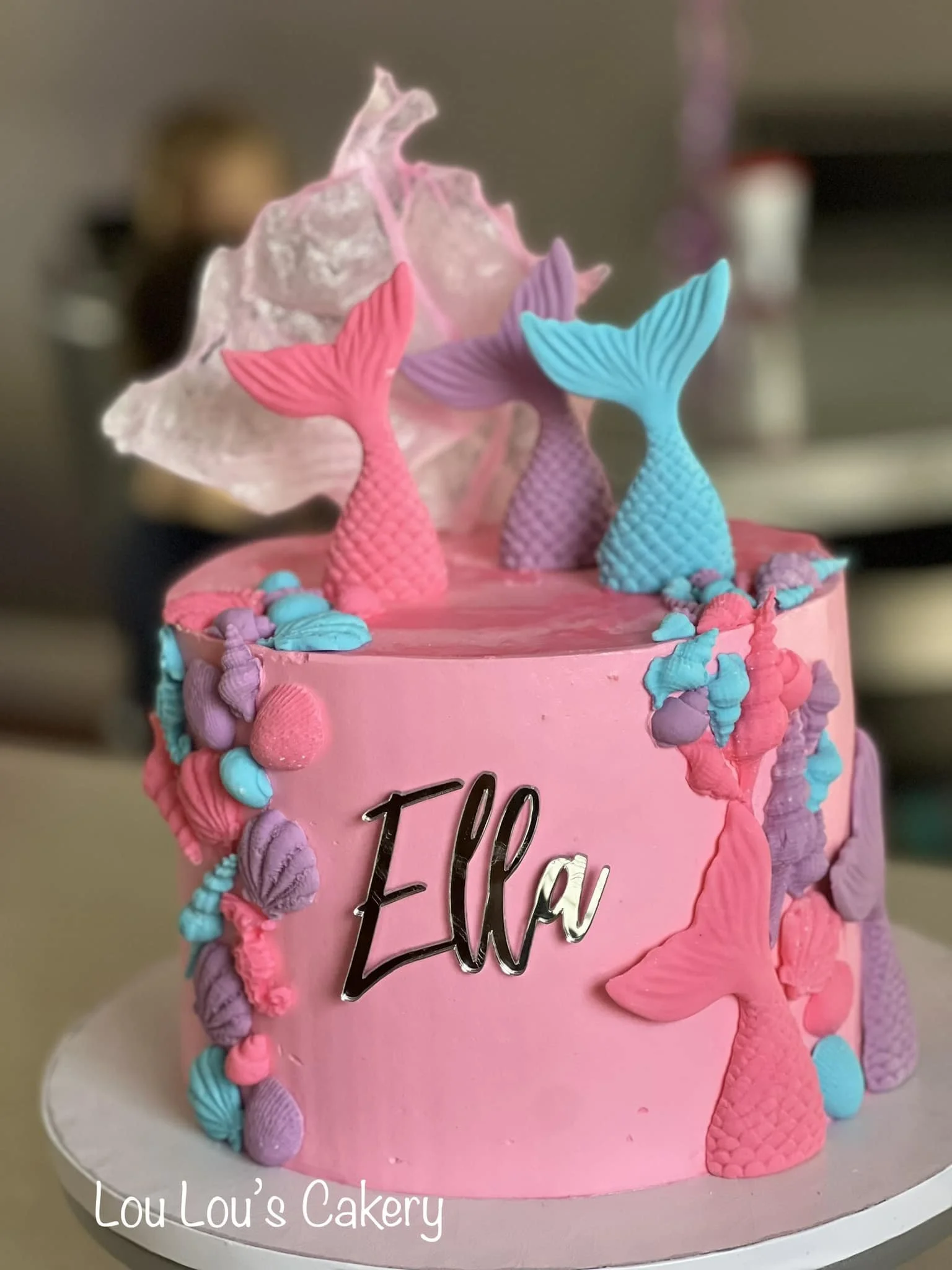 Pink birthday cake with mermaid tail decorations and shell accents, with the name 'Ella' written on the side.