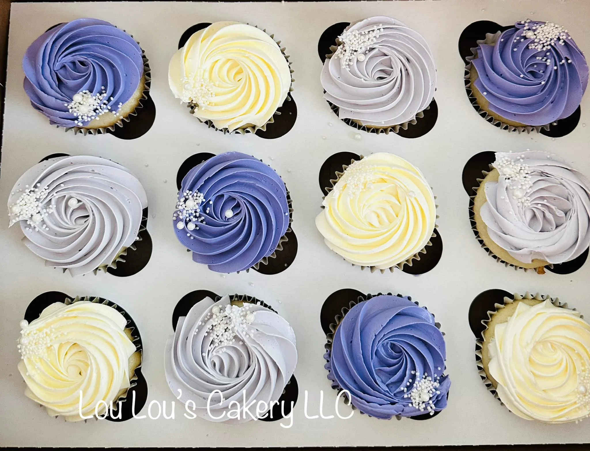 A tray of twelve cupcakes with swirled frosting in blue, white, and light purple, decorated with small white sugar pearls, from Lou Lou's Cakery LLC.