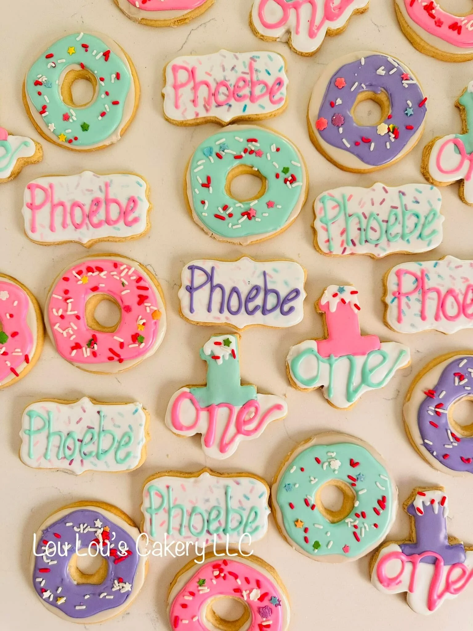 Assorted decorated cookies with words 'one' and 'Phoebe' and colorful sprinkles, arranged on a surface.