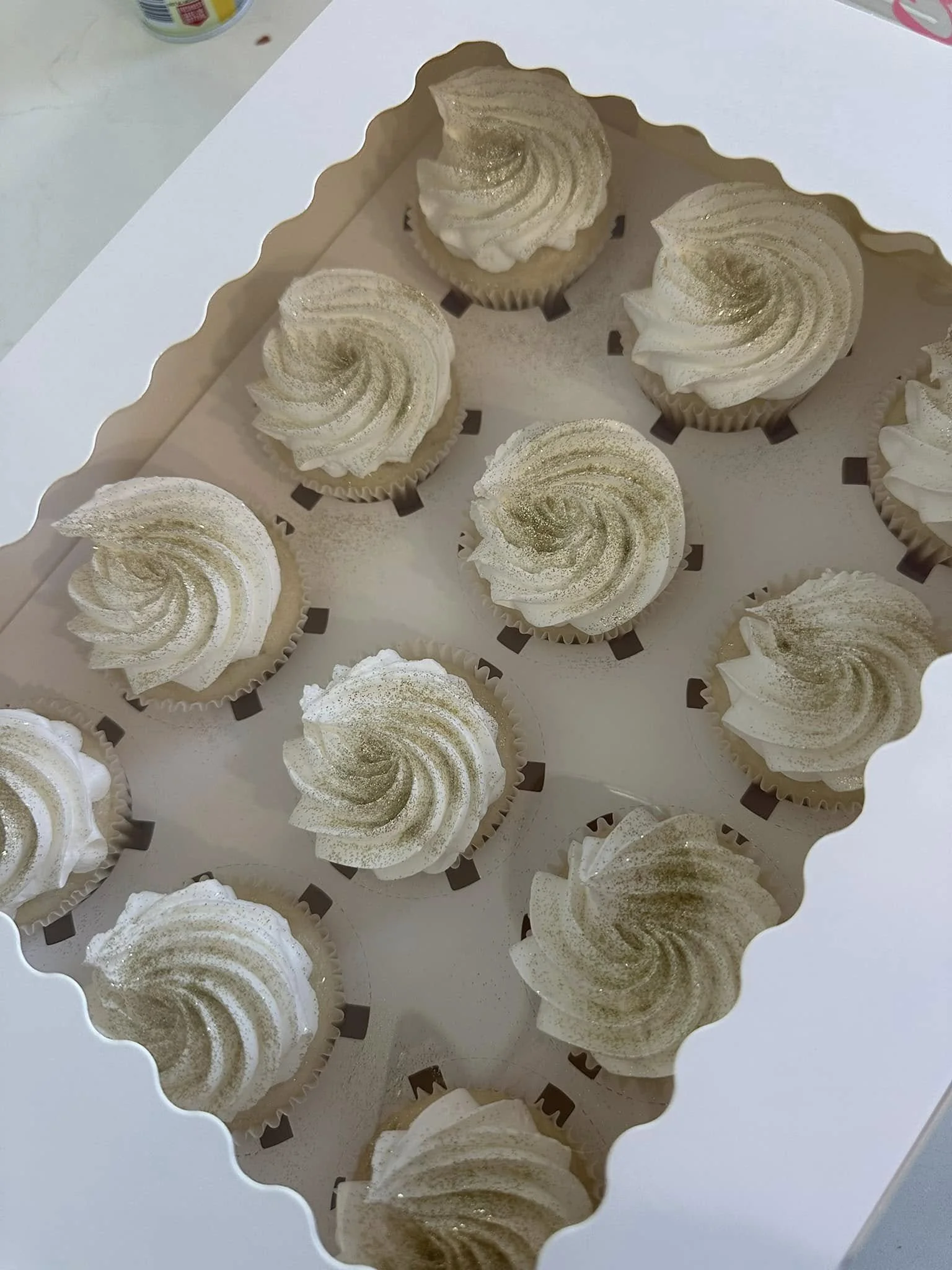 A white box containing twelve cupcakes with white swirled frosting and gold glitter topping.