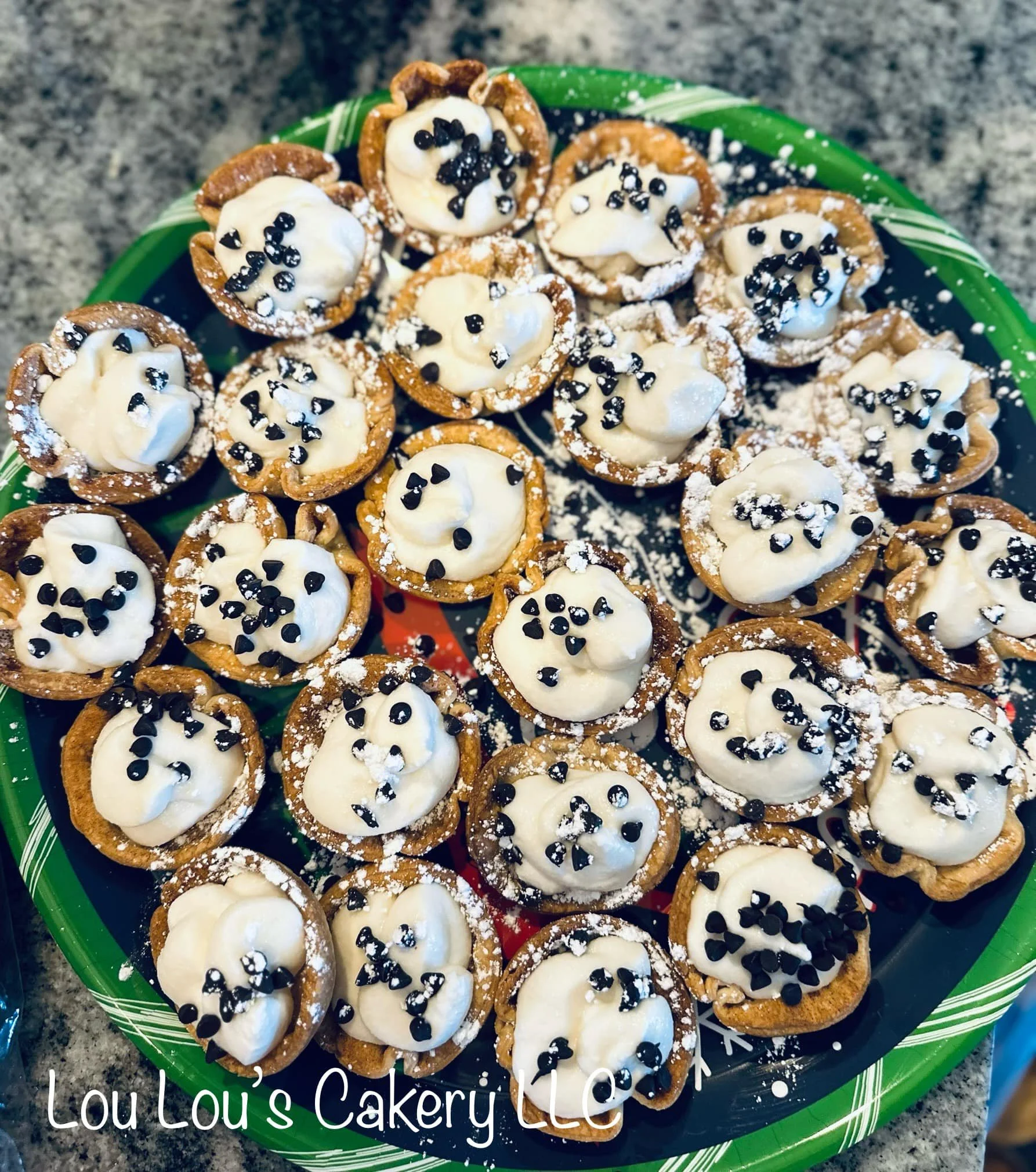 Mini tartlets filled with creamy filling, topped with chocolate sprinkles, arranged on a colorful green and black plate with powdered sugar dusted on top.
