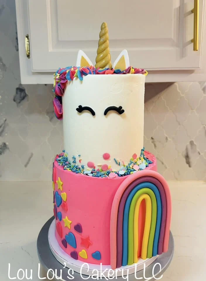 A two-tiered unicorn-themed birthday cake with colorful decorations, including a rainbow on the bottom tier, a unicorn face on the top tier, a unicorn horn and ears, and rainbow sprinkles.