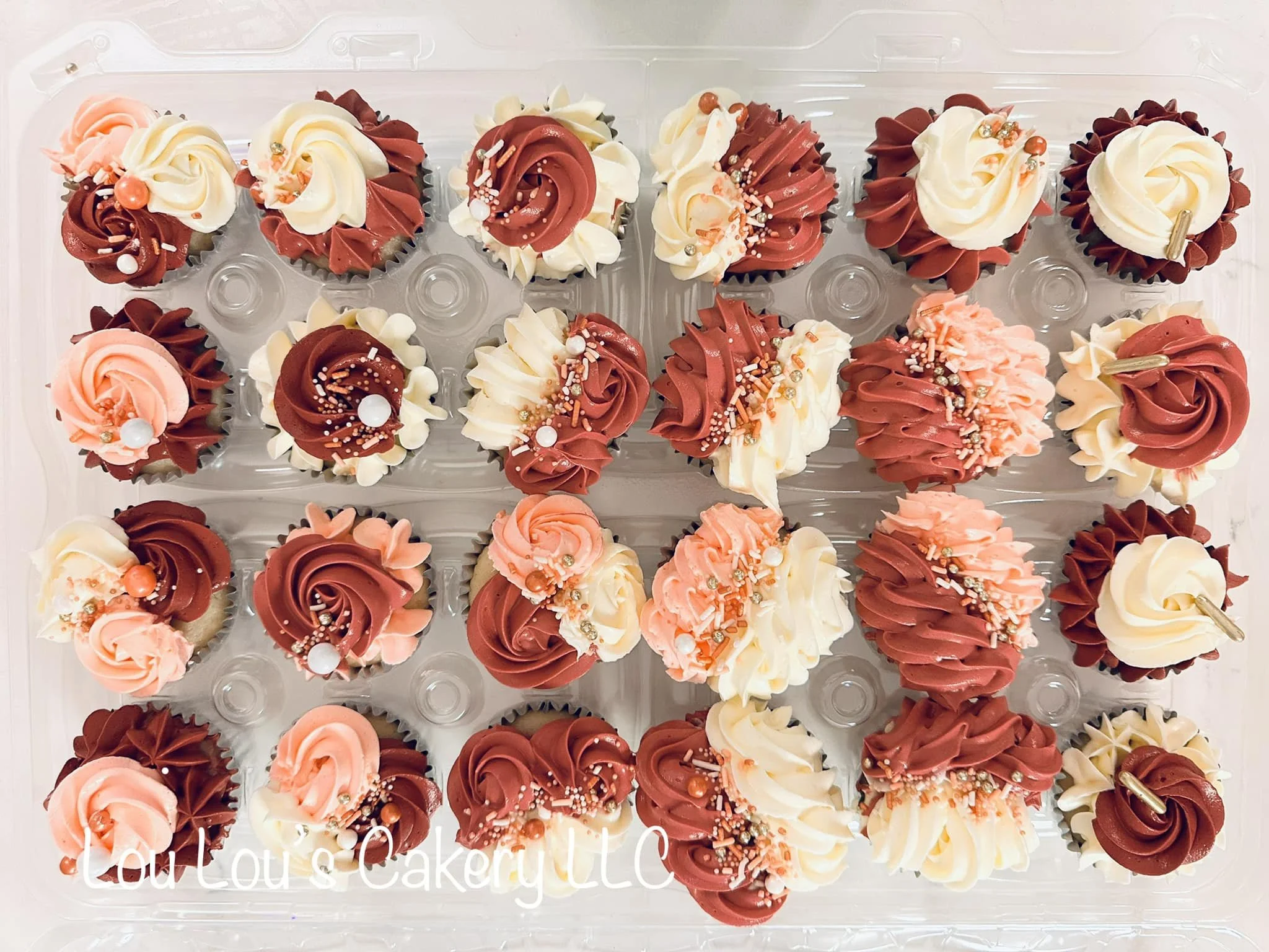 Tray of cupcakes decorated with pink, white, and chocolate-colored frosting, topped with sprinkles and small decorative items.