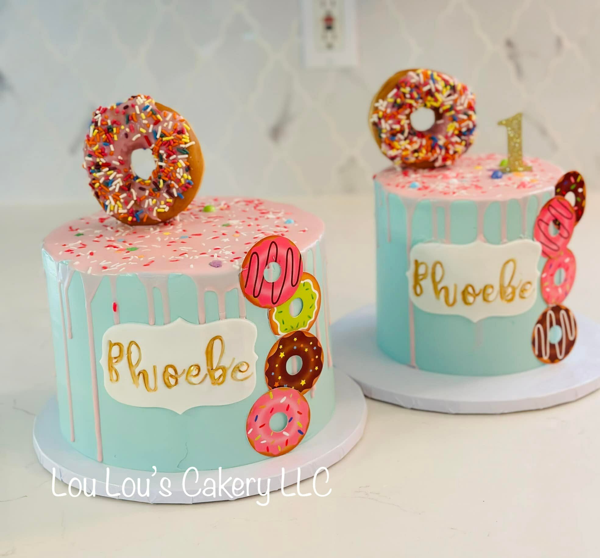 Two birthday cakes with pink and blue drip icing, decorated with colorful sprinkles and topped with a donut-shaped cake decoration. Each cake has a gold number one candle and a white fondant plaque with the name 'Phoebe' written in gold. One cake has