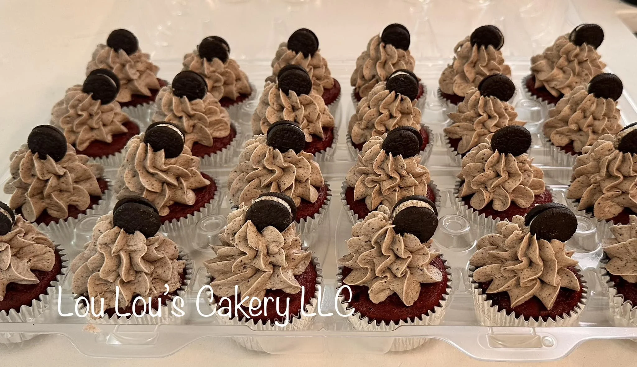 Multiple red velvet cupcakes topped with chocolate frosting and a small Oreo cookie on a clear plastic tray.