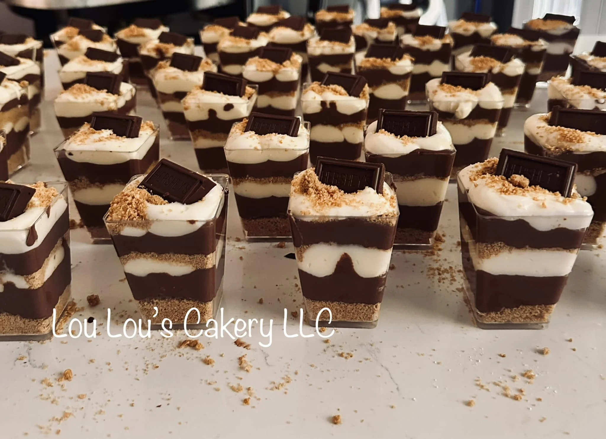Several individual dessert parfaits in clear cups, layered with chocolate, whipped cream, and crumbs, topped with a chocolate piece and crumbs, labeled Lou Lou's Cakery LLC.