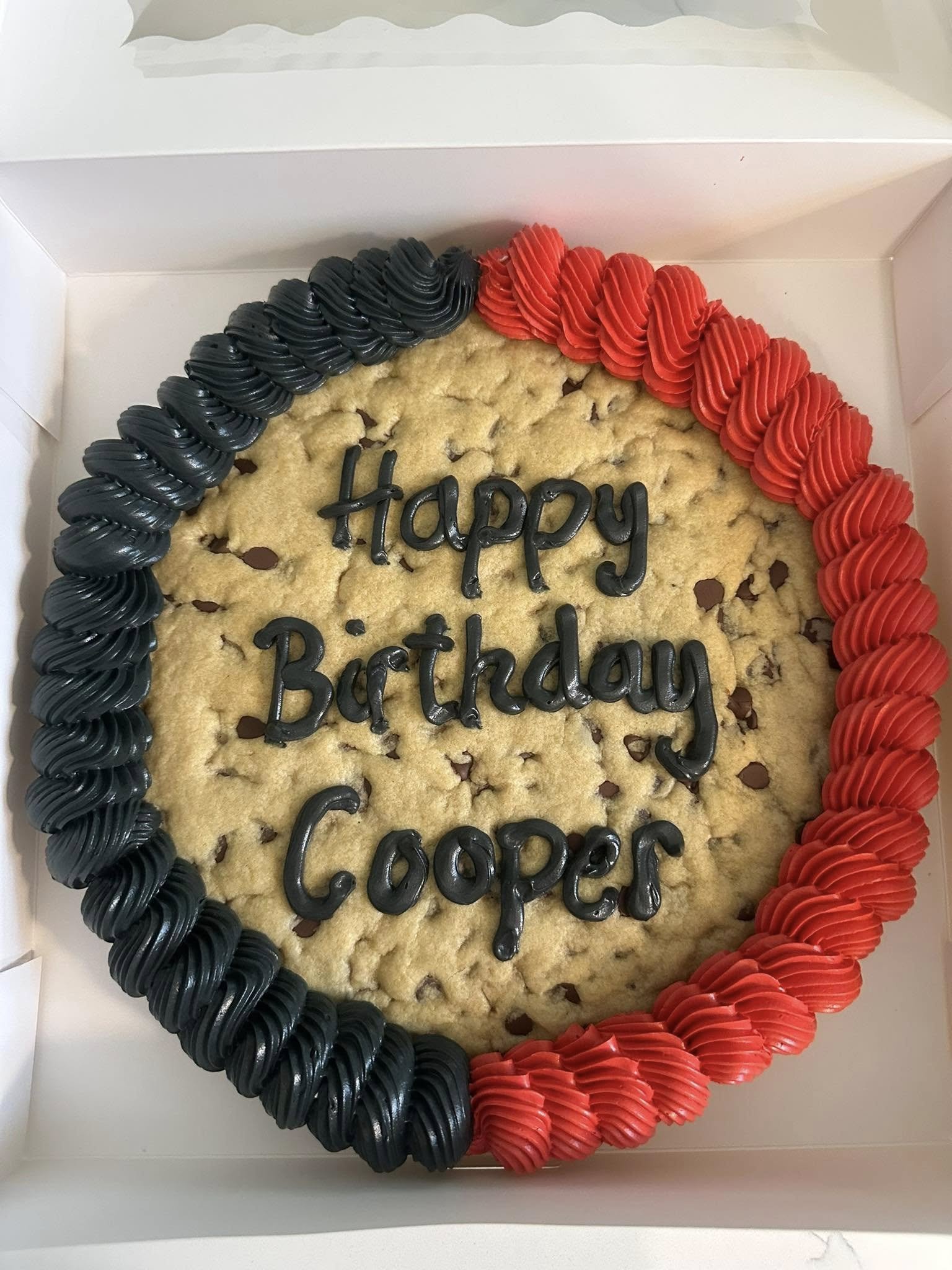 A cookie cake in a box with black and red piped icing, decorated with the message, "Happy Birthday Coopers" written in black icing.
