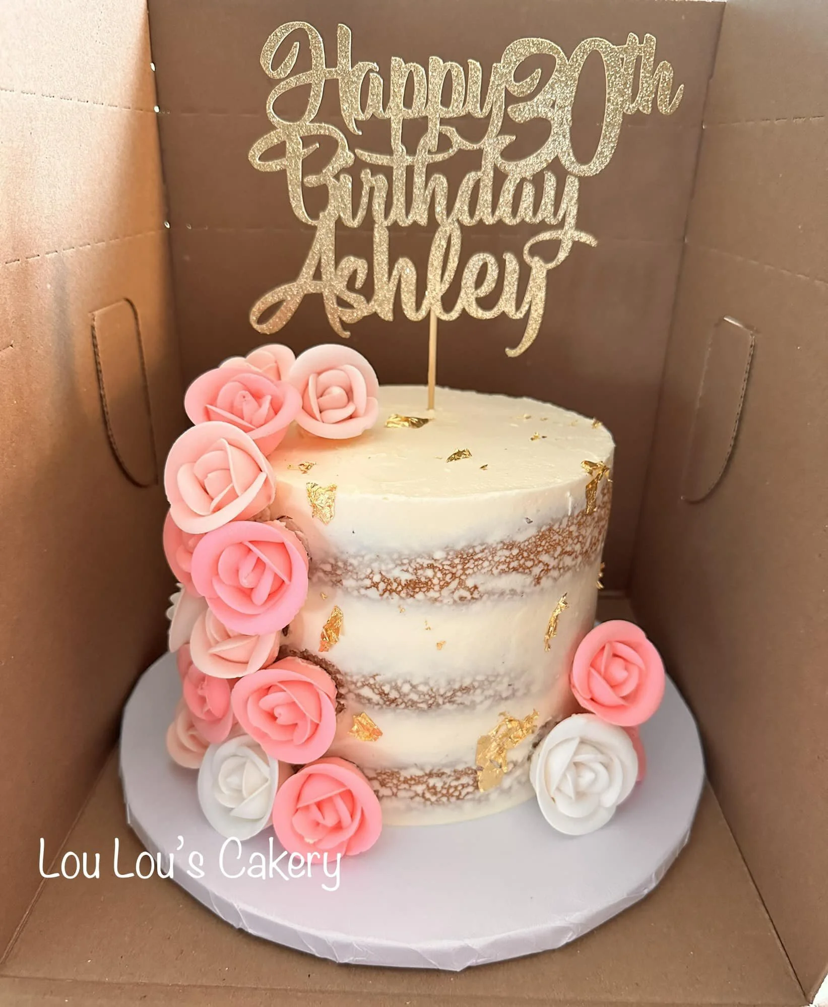 A layered birthday cake decorated with pink and white roses, gold leaf accents, and a glittery gold "Happy 30th Birthday Ashley" topper inside a brown gift box.