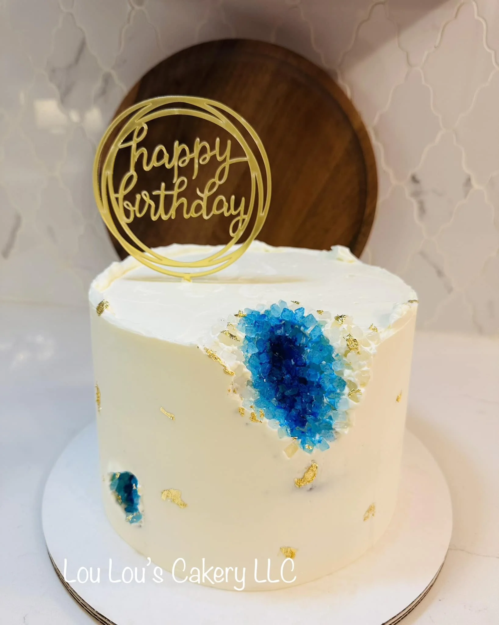 A white birthday cake with blue crystallized decoration, gold leaf accents, and a gold "happy birthday" topper, placed on a white surface with a wooden and decorative background.