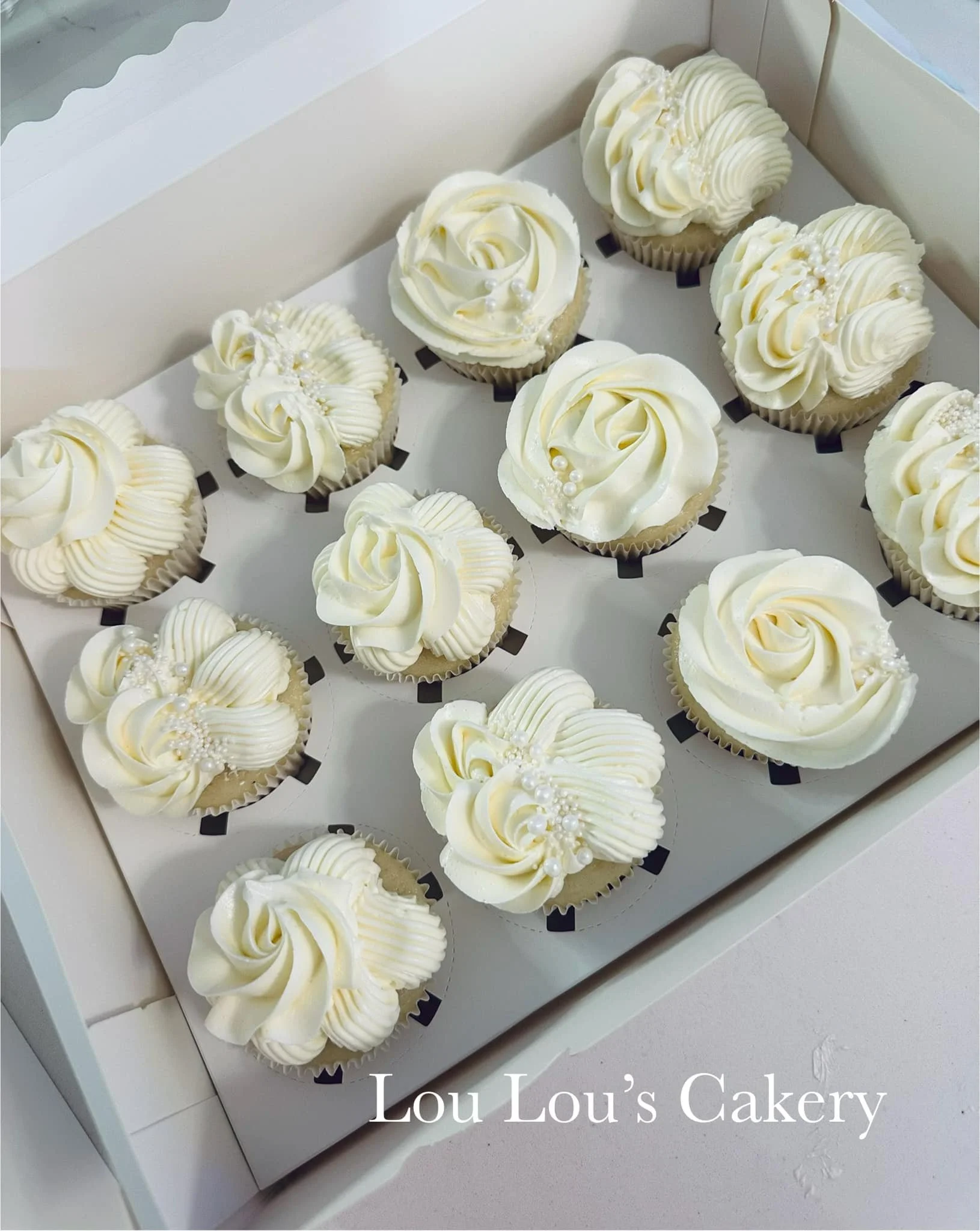 A box of white cupcakes decorated with white frosting and pearl-like sprinkles, labeled 'Lou Lou's Cakery'.