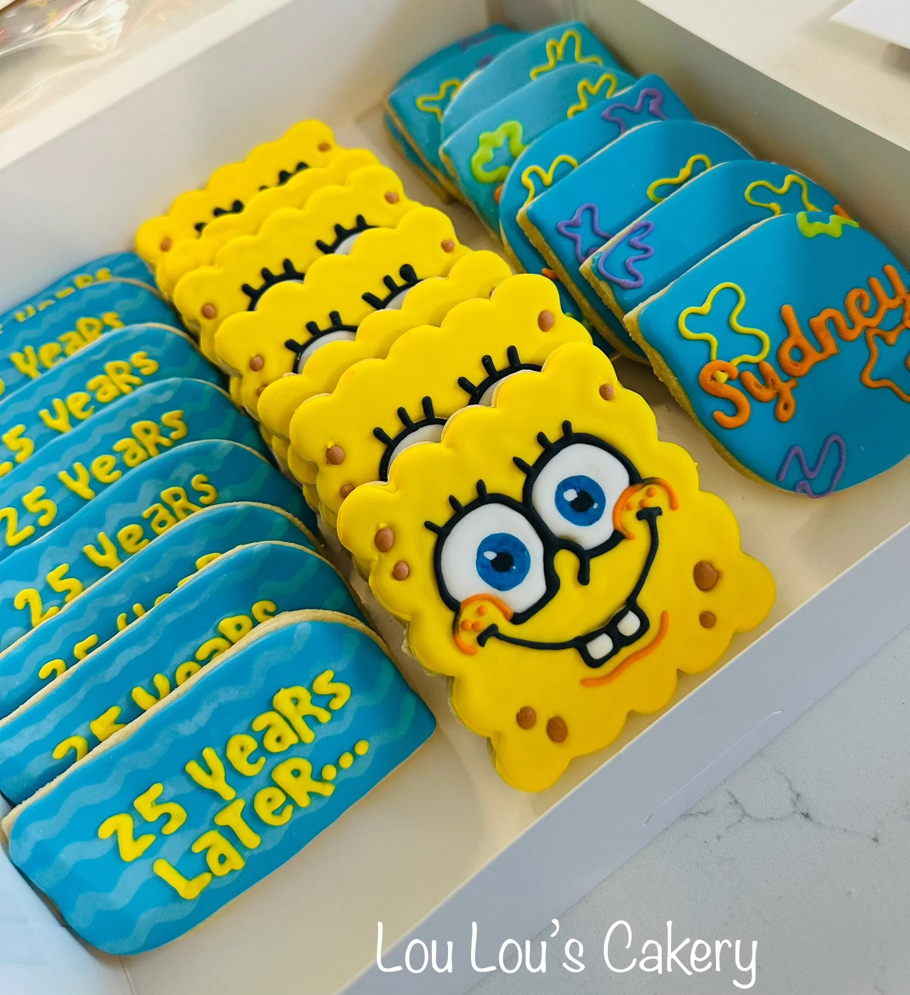 Decorated cookies featuring a SpongeBob SquarePants face, cupcakes with the message '25 Years Later...', and blue cookies with colorful designs, in a white box from Lou Lou's Cakery.