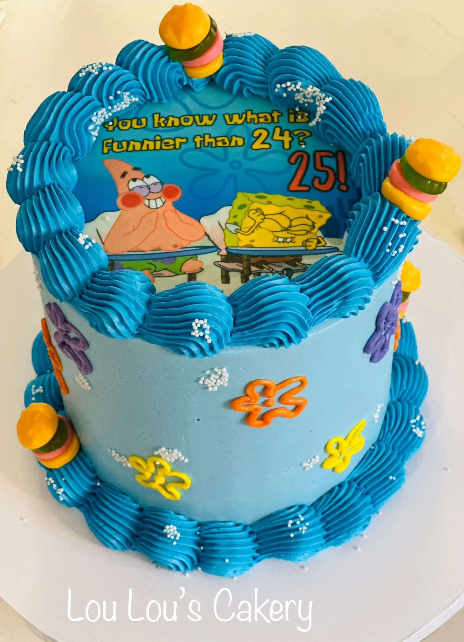 A two-tier blue birthday cake decorated with blue frosting piped around the edges, colorful butterfly and flower decorations, and small burger and fries toppers. The top of the cake features an edible image of SpongeBob SquarePants and Patrick Star w
