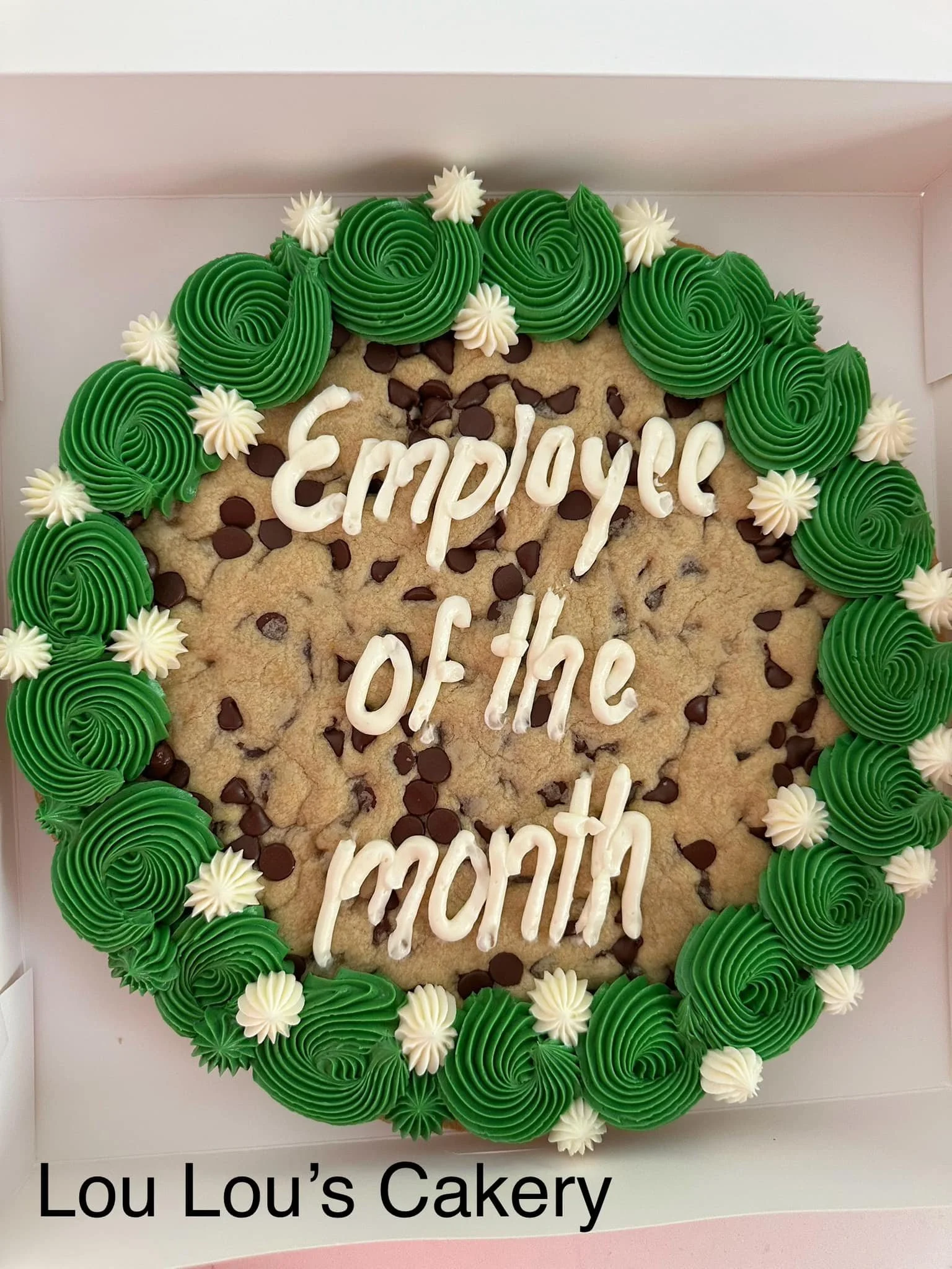 A large cookie decorated with green and white icing around the edges, with chocolate chips on top, and the words 'Employer of the month' written in white icing in the center. The cookie is in a box with the text 'Lou Lou's Cakery' inside.