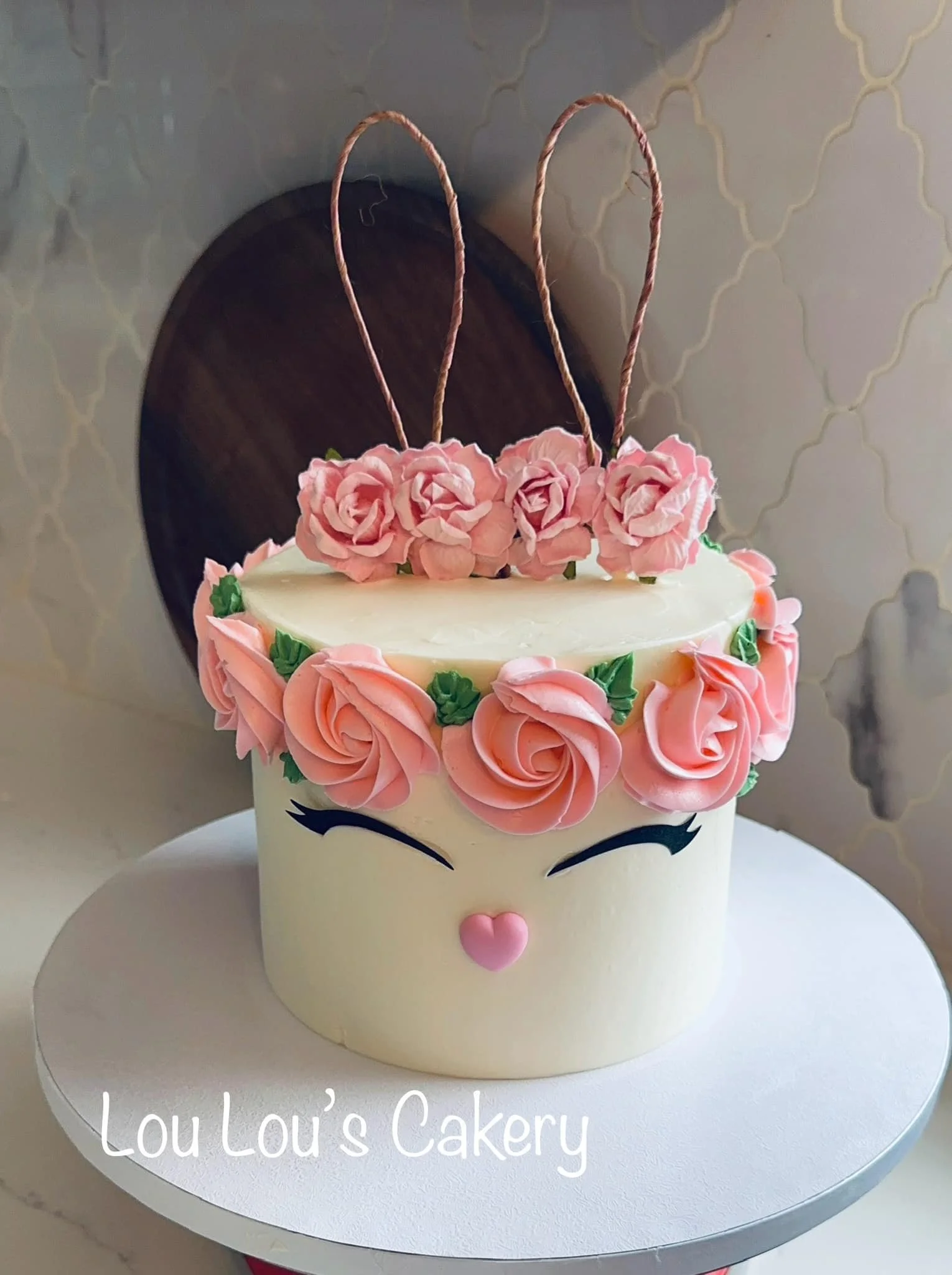 A decorated cake with a unicorn face, pink floral decorations, and two braided topping sticks. The cake has closed eyes with long eyelashes and a small pink heart-shaped nose. It is placed on a white cake board with the text "Lou Lou's Cakery" writte