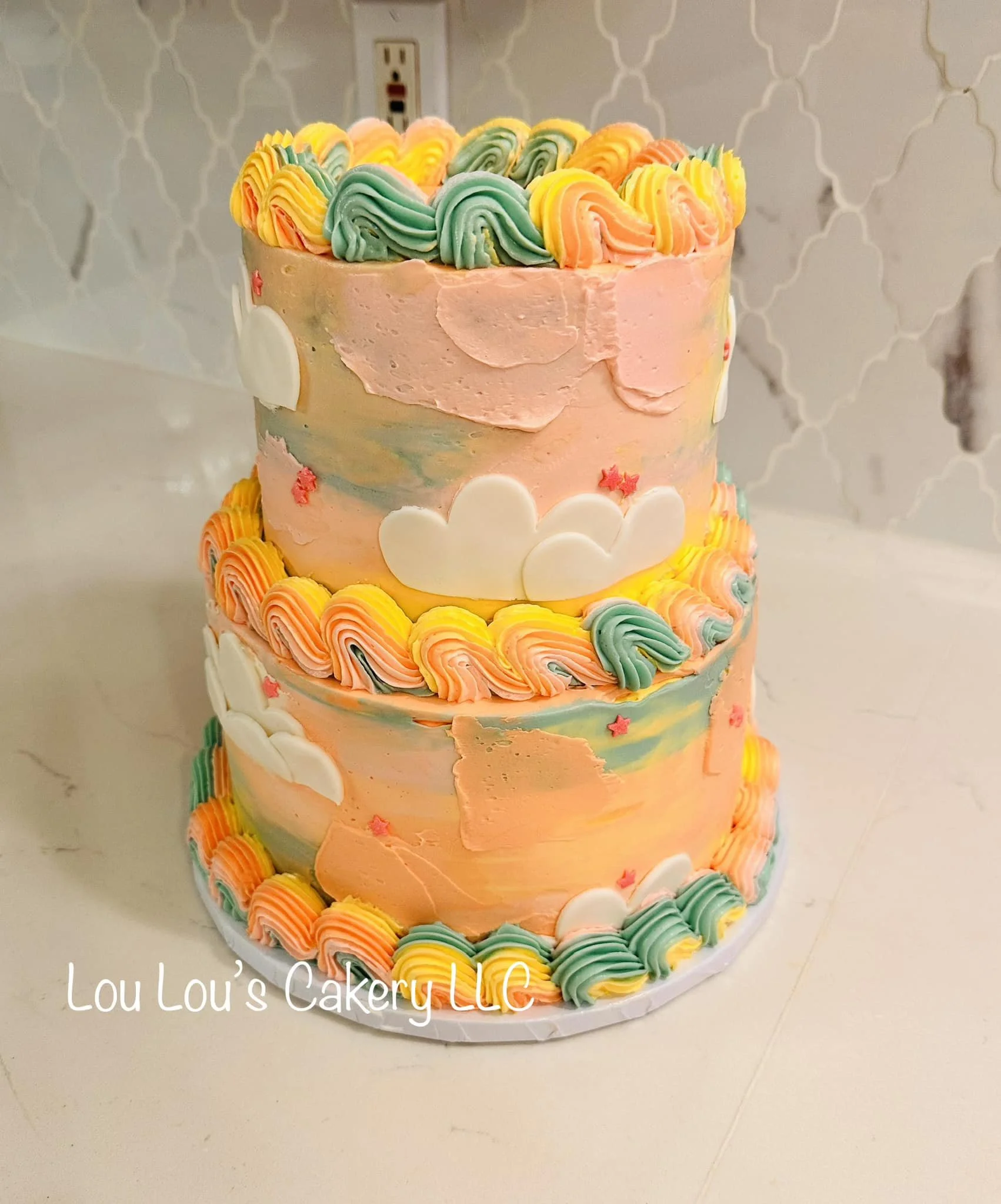 Two-tiered pastel-colored cake with colorful piped frosting, white cloud-shaped decorations, and small star and heart accents, placed on a white surface in front of a patterned wall and electrical outlet.