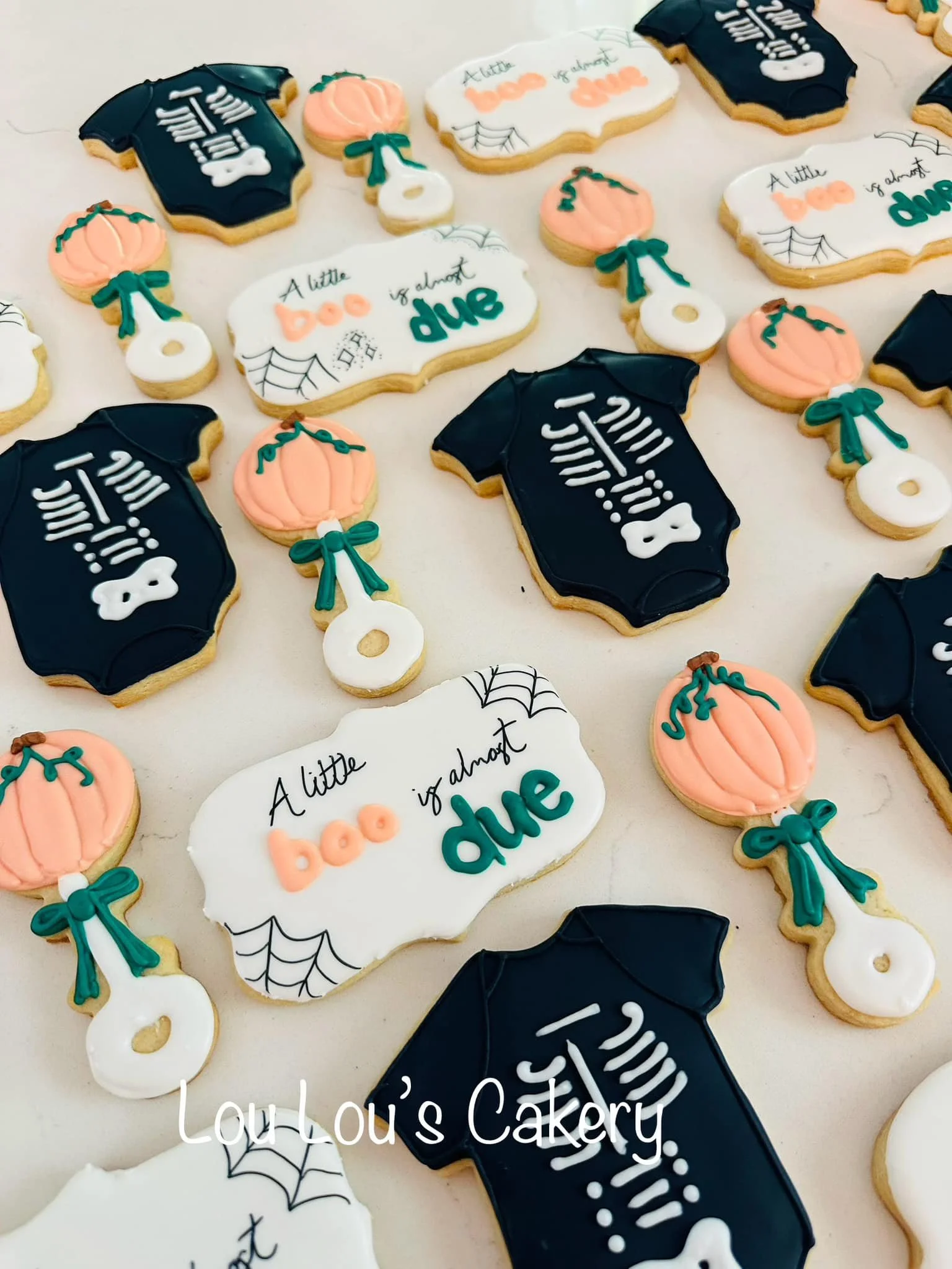 Halloween-themed decorated sugar cookies including skeleton bodysuits, pumpkins with bows, and "A little boo of almost due" signs, on a white surface, from Lou Lou's Cakery.