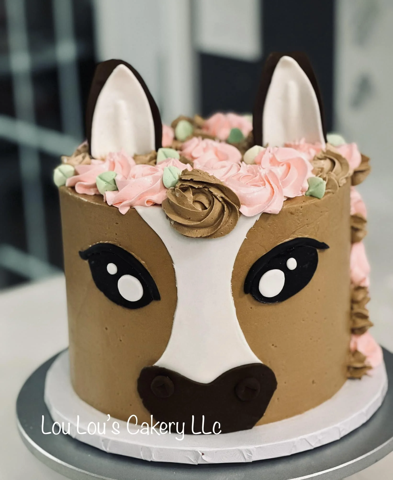A cake decorated to resemble a horse's face with chocolate, pink, and white toppings, featuring ears, eyes, and a nose, with pastel-colored flower and swirl decorations on top.