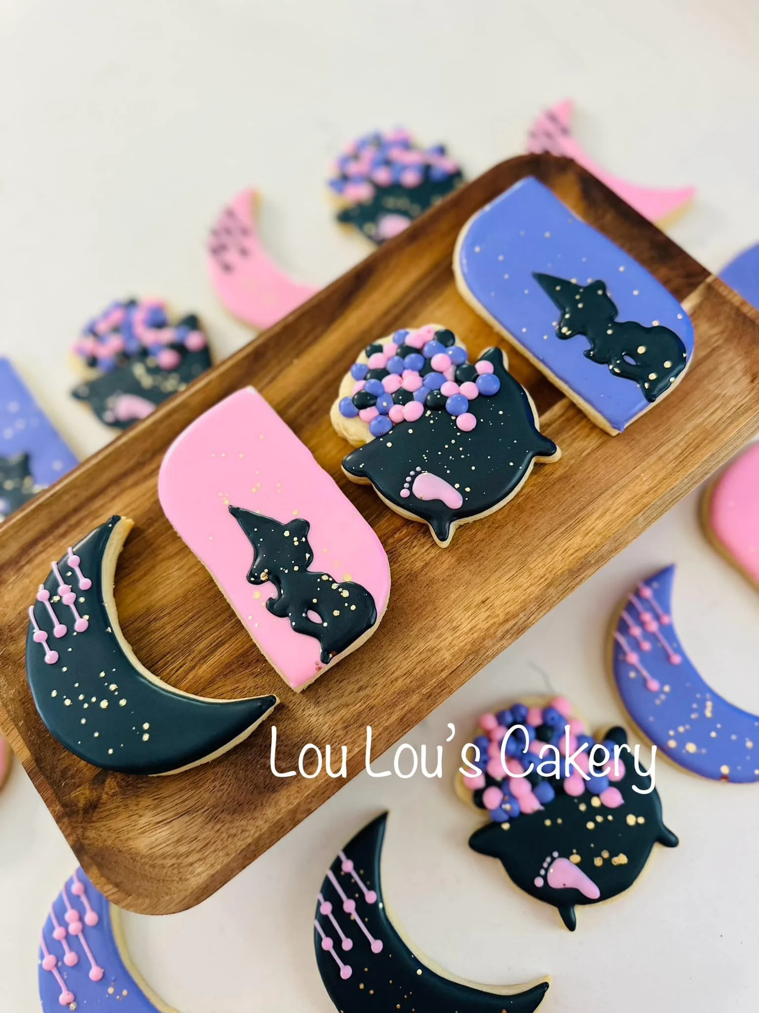 Decorative cookies with moon and unicorn shapes, decorated with pastel pink, purple, and black icing, placed on a wooden tray and white surface, with the text "Lou Lou's Cakery" visible.