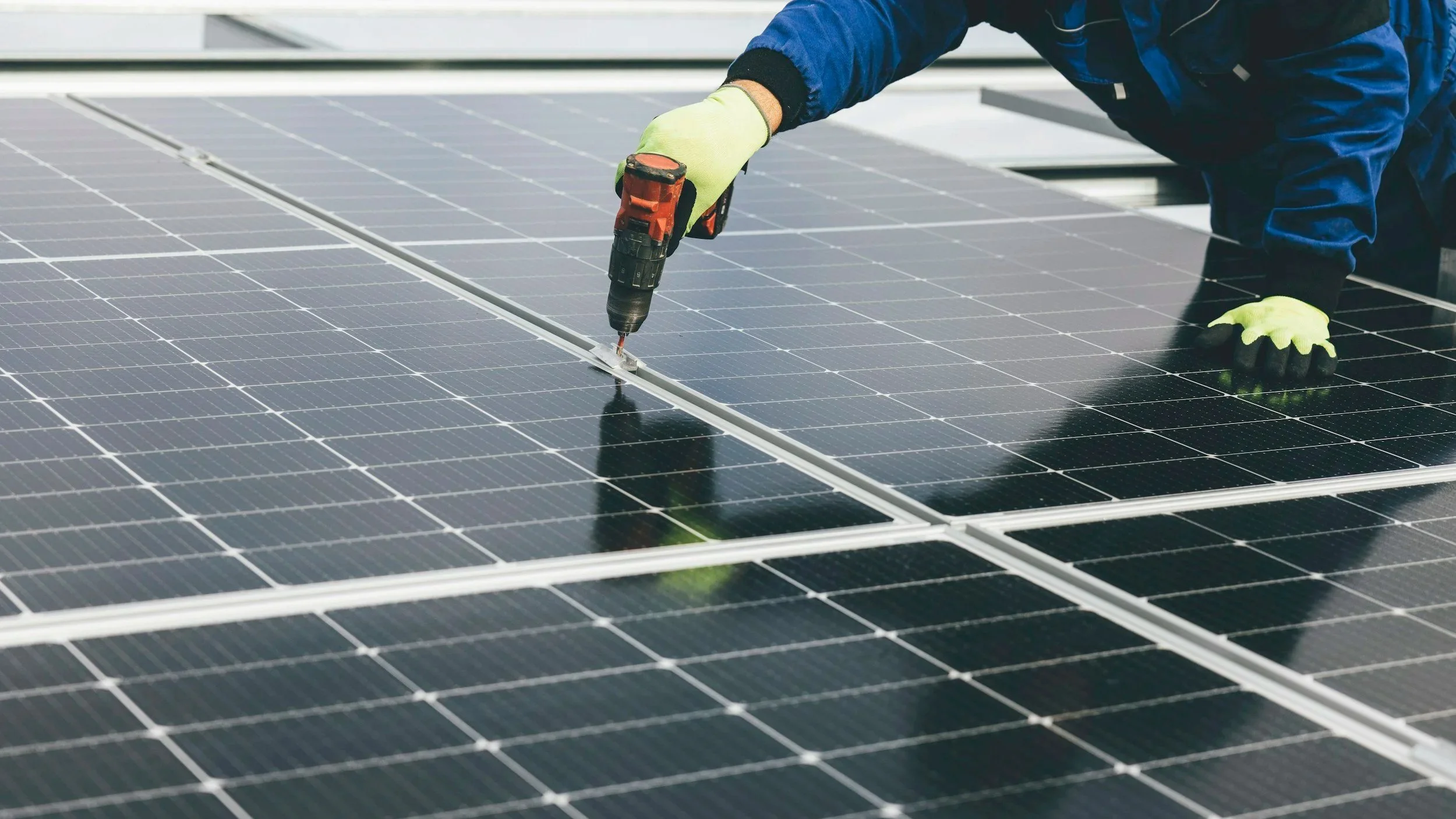 A person installing or repairing solar panels on a rooftop using a cordless drill, wearing gloves and a blue jacket.