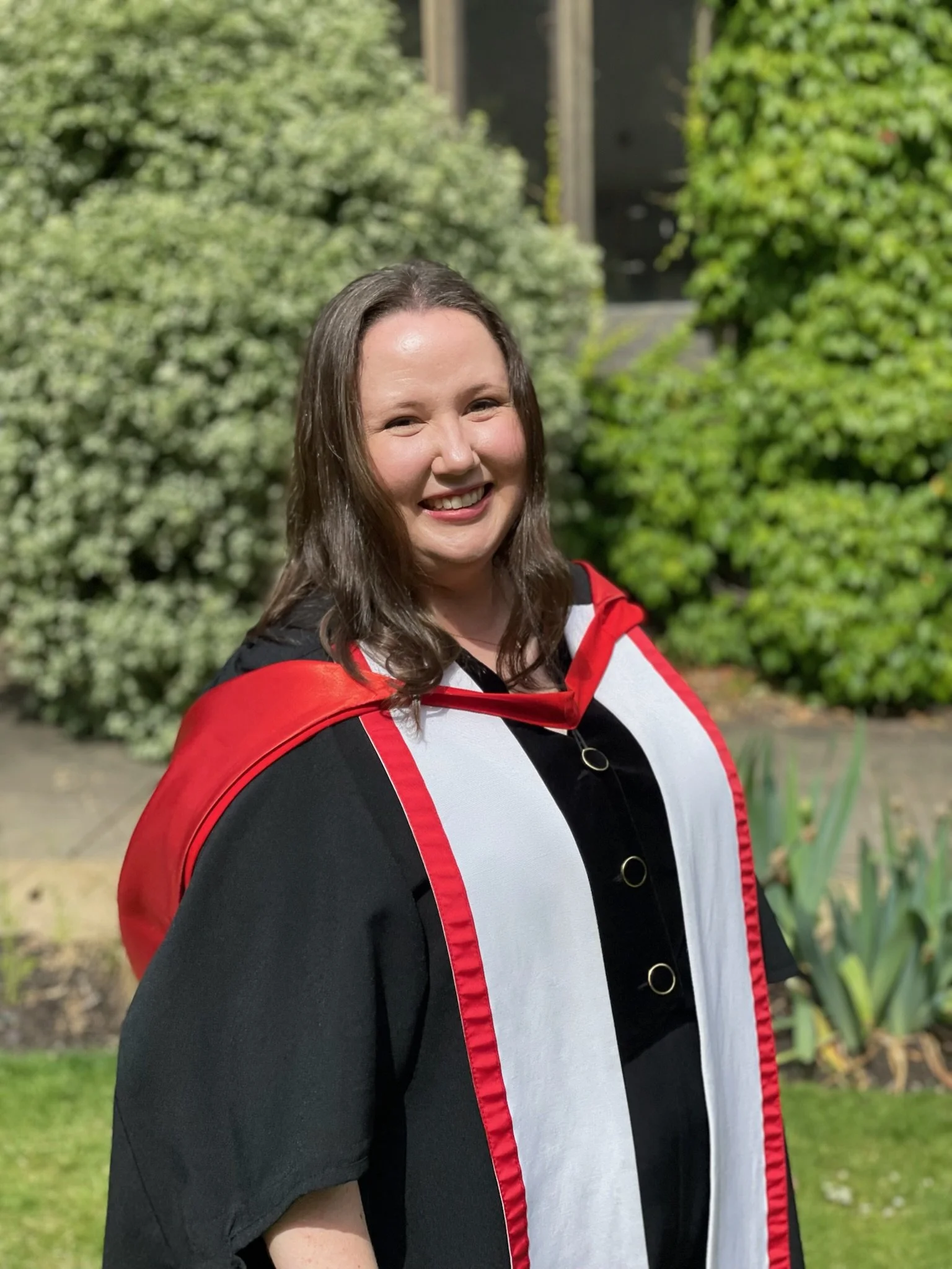 Smiling woman with shoulder-length brown hair, standing outdoors near green foliage.