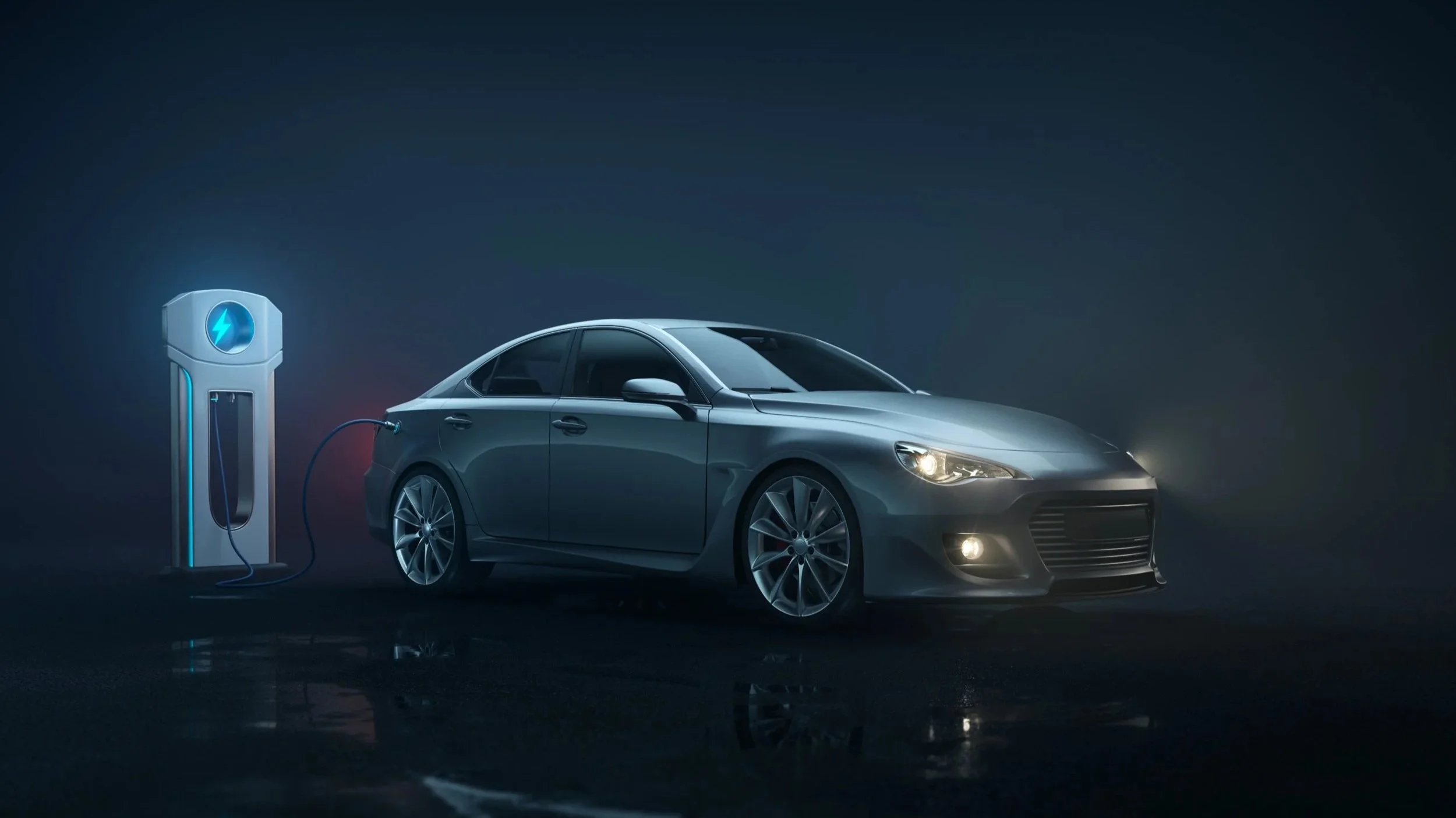 A silver electric car connected to a charging station in a dark setting, with the car's headlights on.