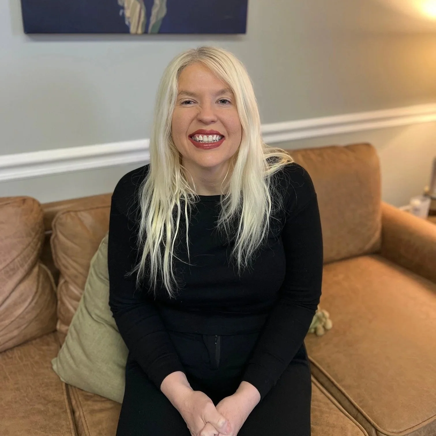 A woman with long blonde hair, wearing a black long-sleeve shirt, sitting on a beige couch, smiling at the camera.