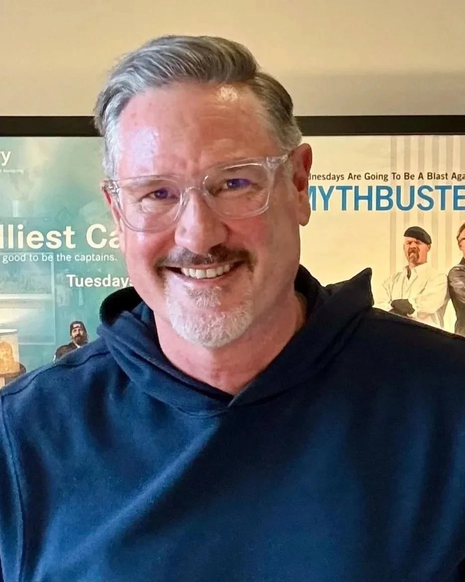 A smiling man with gray hair and glasses standing in front of a poster with the words "MYTHBUSTERS" and images of two men wearing white coats.