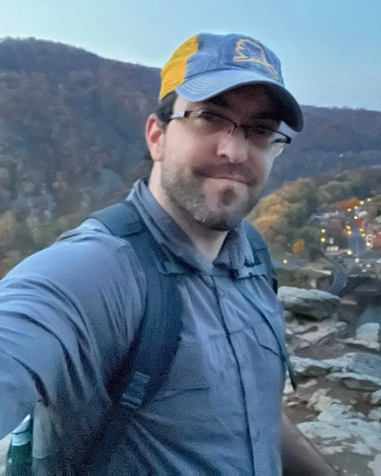 Man with brown beard wearing semi rimless glasses, a blue and yellow ball cap and a blue button down with a mountain scene in the background
