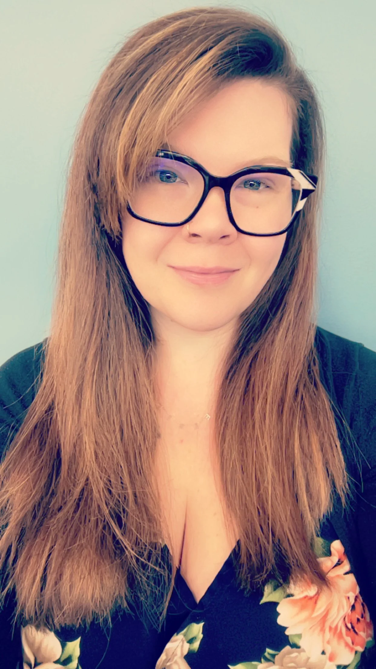 young woman with below shoulder length light brown hair wearing large black glasses with a black sweater over a navy blue shirt with pink and green roses