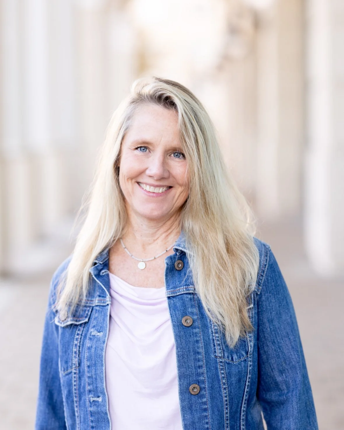 A woman with long blonde hair smiling outdoors, wearing a pink shirt and layered necklaces.