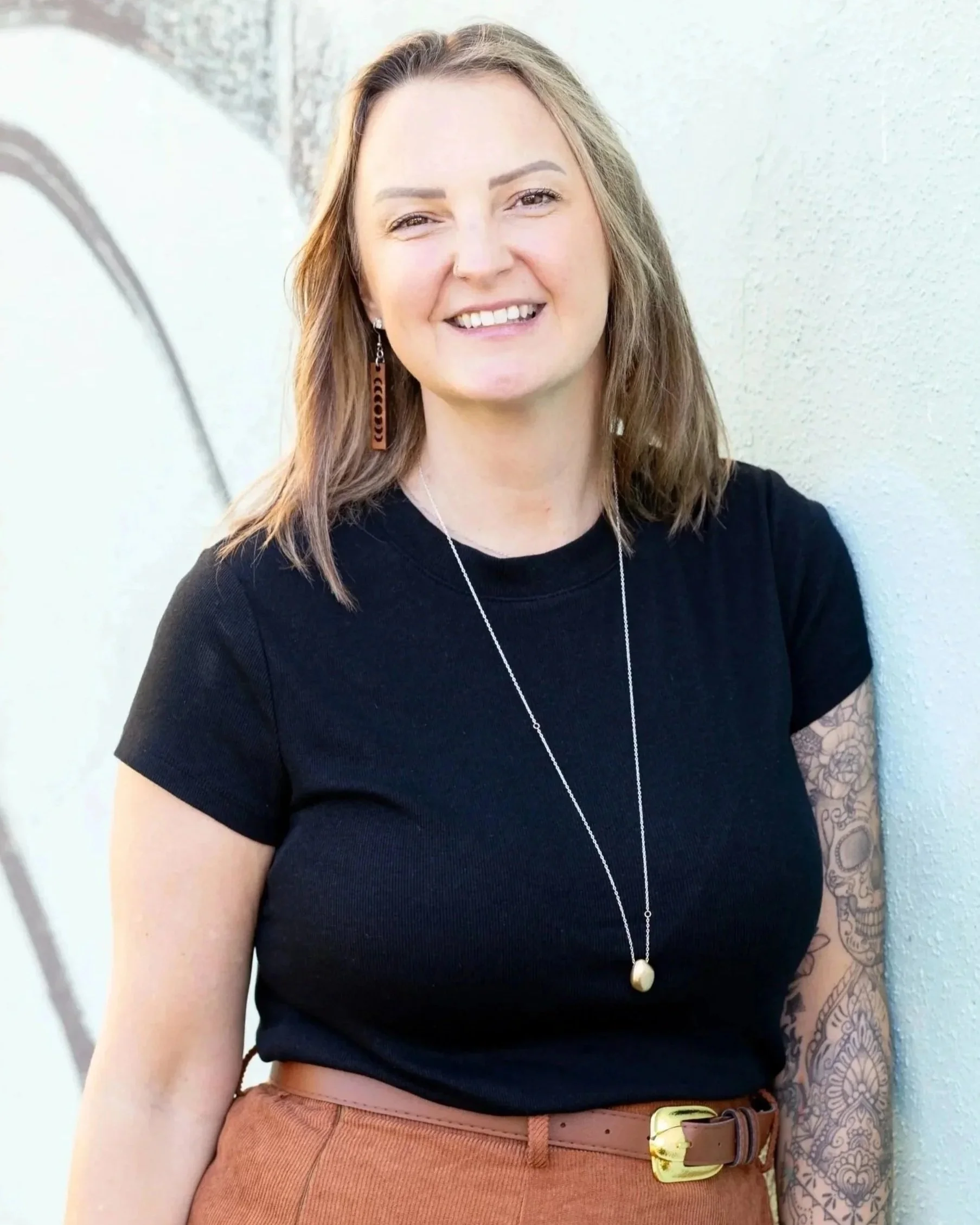 A woman with shoulder-length light brown hair, smiling, wearing a black T-shirt, orange pants, a long necklace, and a tattoo sleeve on her right arm, standing against a textured off-white wall with graffiti.