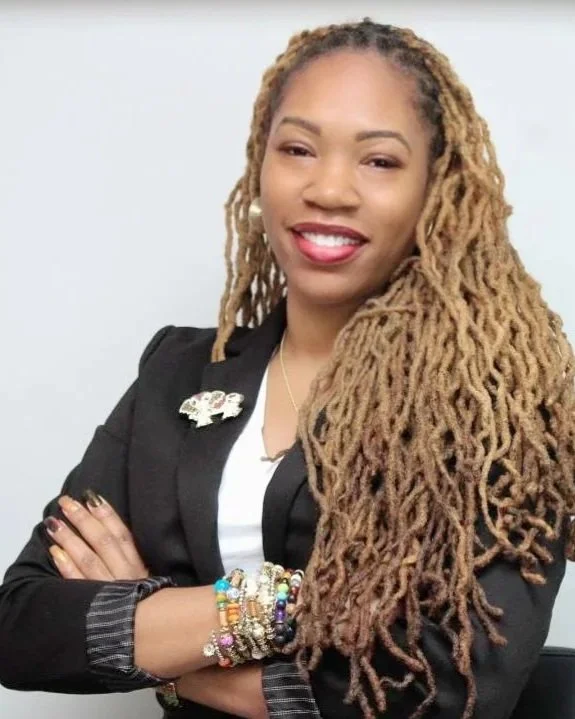 A woman of color, with golden chest length locks smiling in a white top and black blazer with fun beaded bracelets.