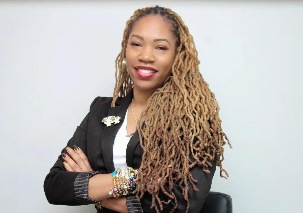 A woman of color, with golden chest length locks smiling in a white top and black blazer with fun beaded bracelets.
