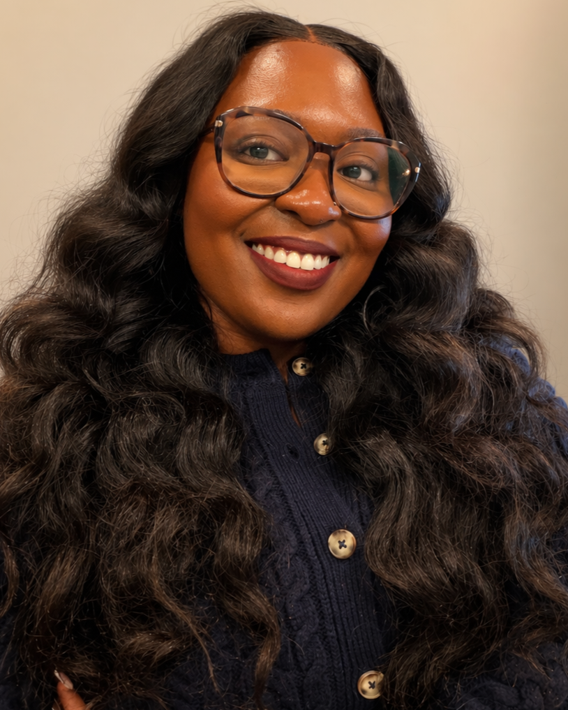 A woman with long, curly dark hair wearing glasses and a dark knitted cardigan, smiling.