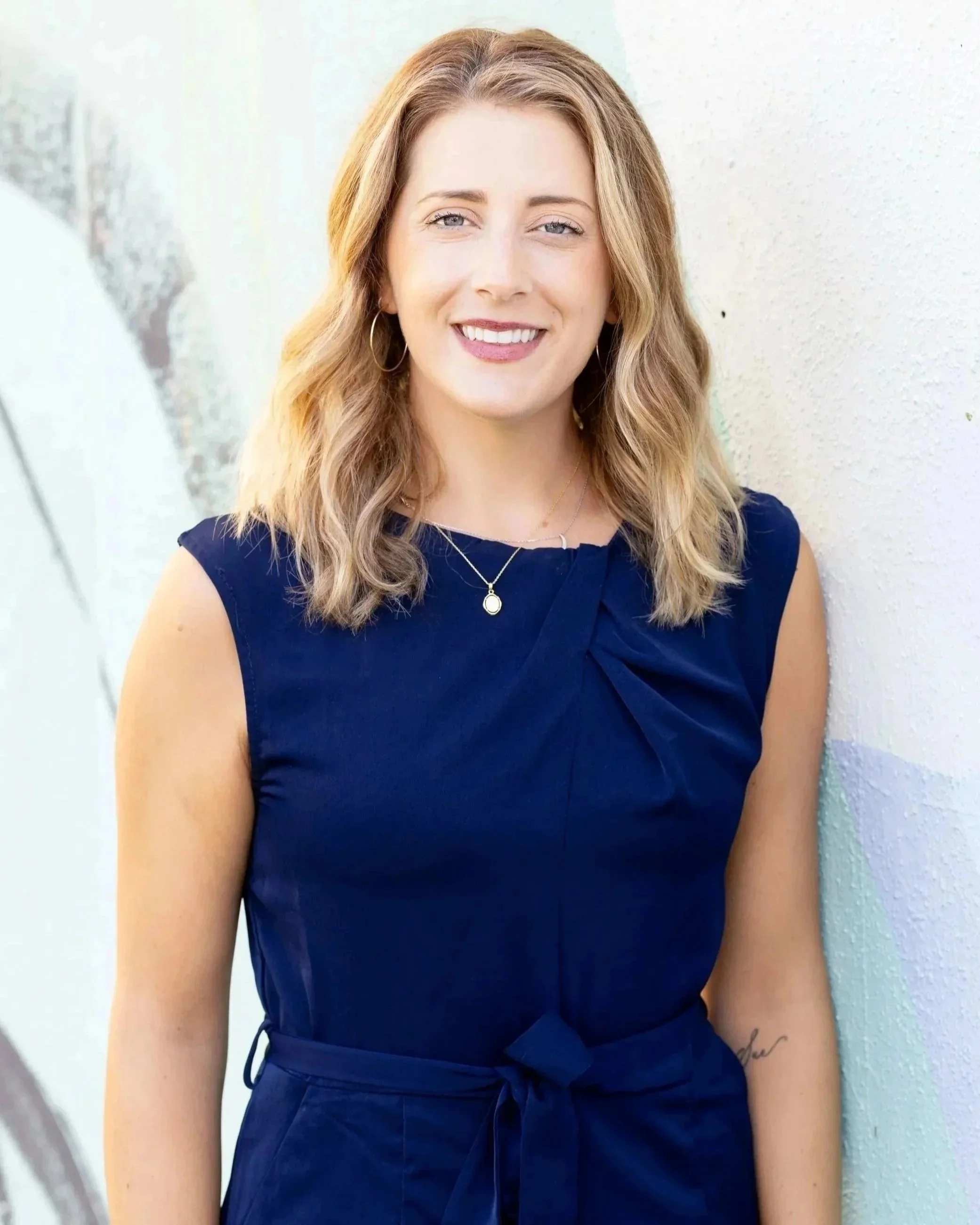 A woman with shoulder-length wavy blonde hair, wearing a navy blue sleeveless dress, gold hoop earrings, a necklace with a pendant, and a tattoo on her left arm, standing against a light-colored textured wall, smiling at the camera.