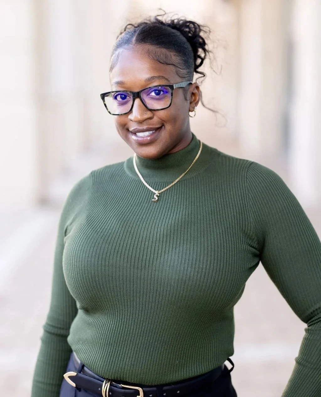 A woman with dark skin wearing a green ribbed turtleneck, glasses, and gold jewelry, smiling at the camera.