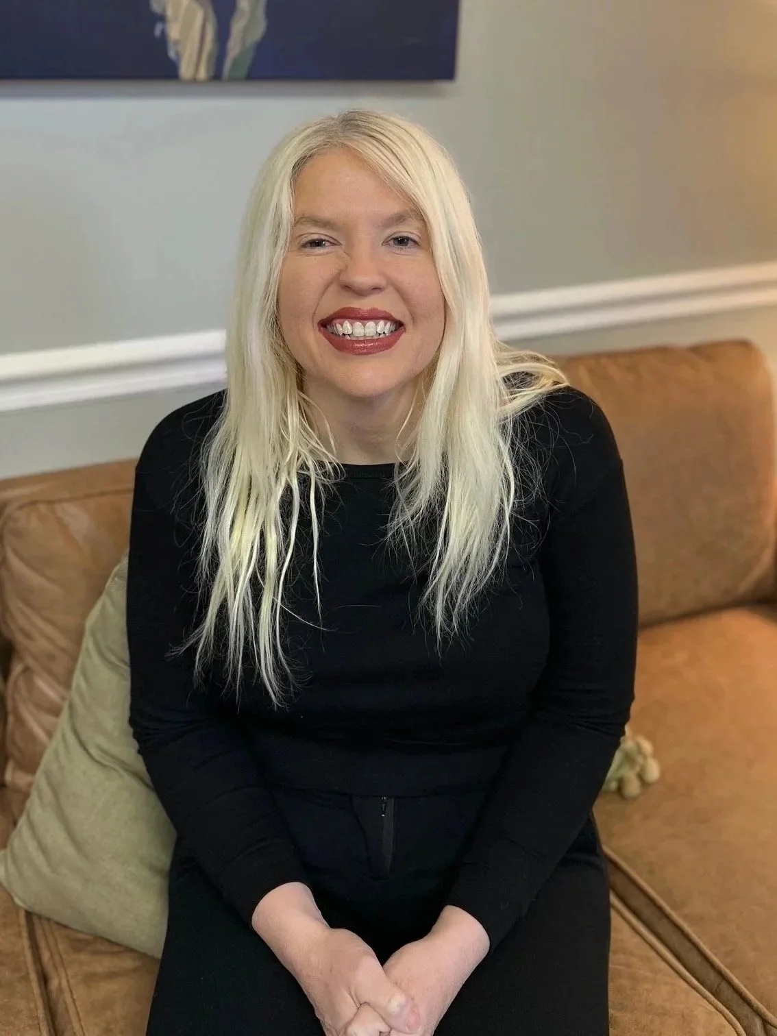 A woman with long blonde hair sitting on a tan couch, smiling with red lipstick, wearing a black long-sleeve top, in a room with light gray walls and artwork in the background.