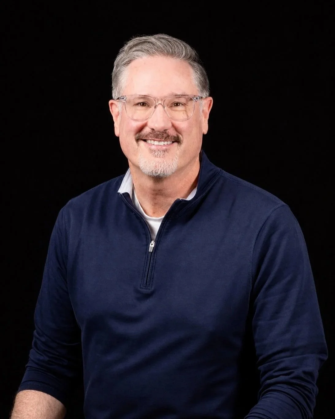 A man with glasses and gray hair smiling in front of a background with books and posters.