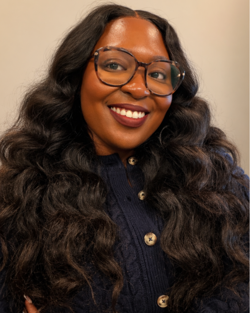 A woman with long, curly dark hair, wearing glasses with tortoiseshell frames, smiling, and dressed in a navy blue button-up top with gold buttons.