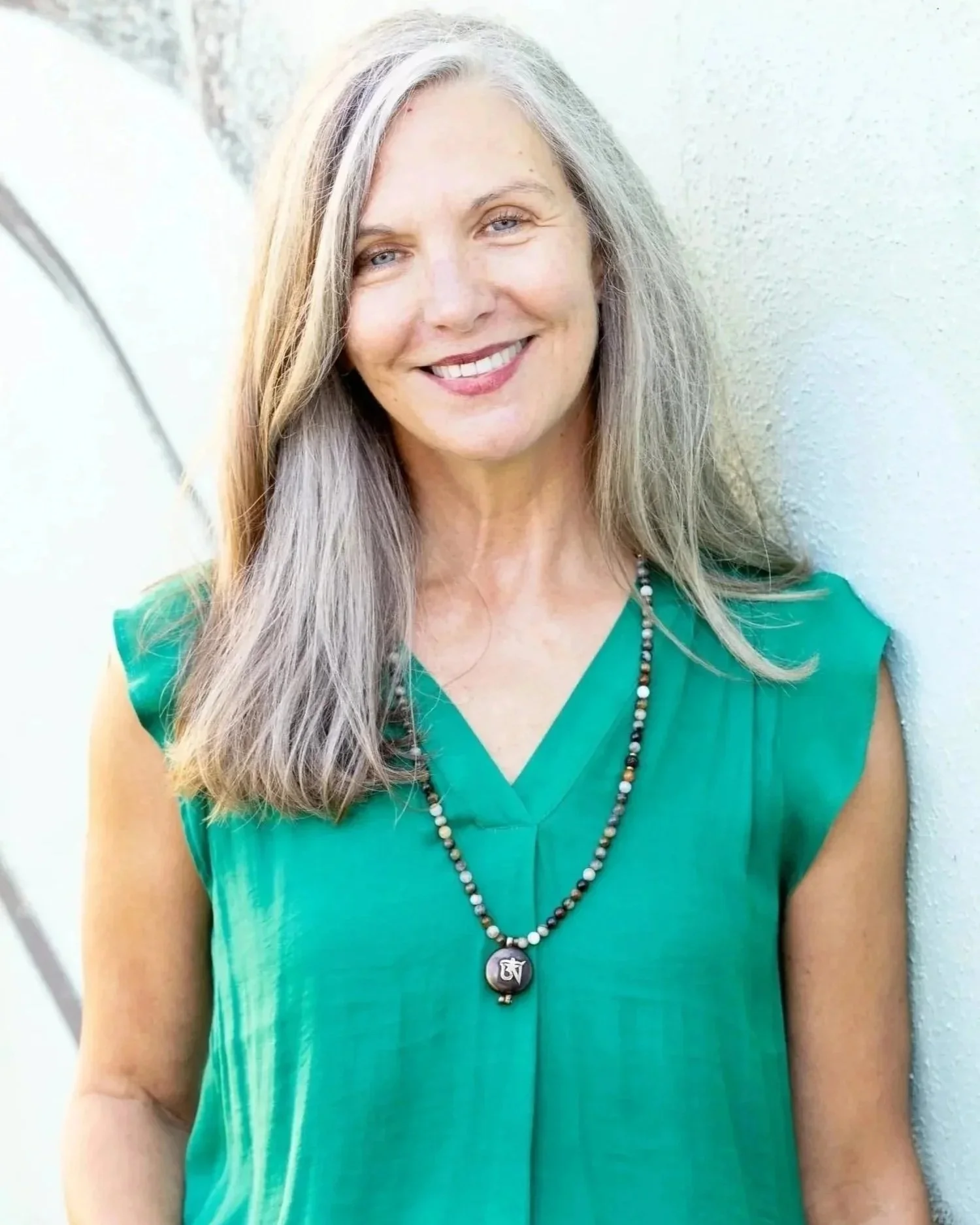 A woman with long gray hair smiling while leaning against a white wall with graffiti.