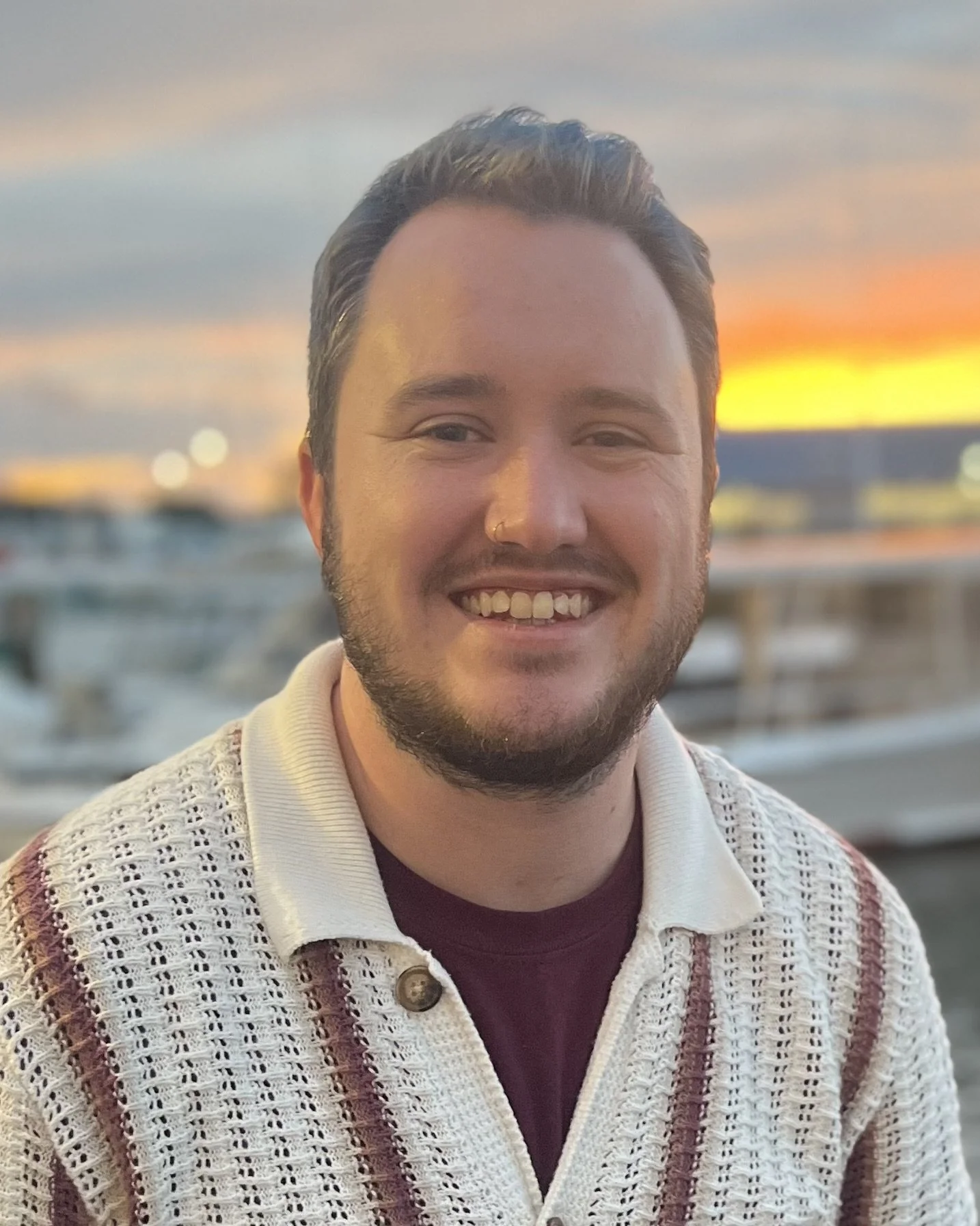 A smiling man with dark hair and a beard, wearing a patterned beige and brown sweater over a maroon shirt, posing outdoors during sunset near water with boats in the background.