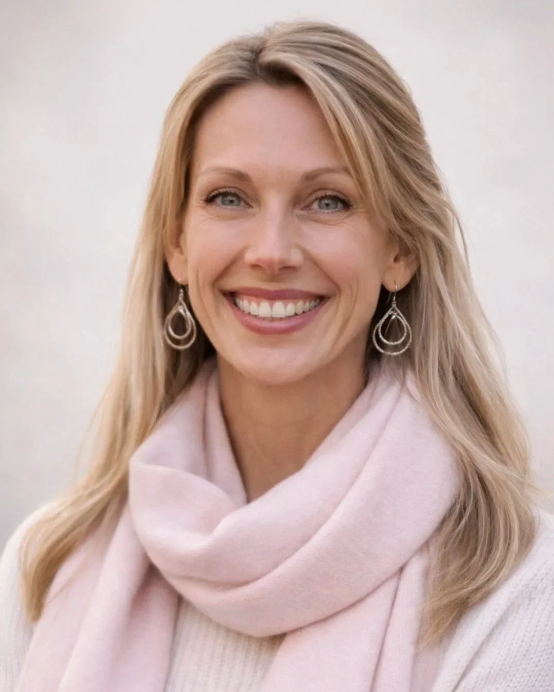Woman with blonde, below the shoulder length hair wearing blue jeans, a white turtleneck shirt with a deep wine colored blazer standing outside under scenic trees